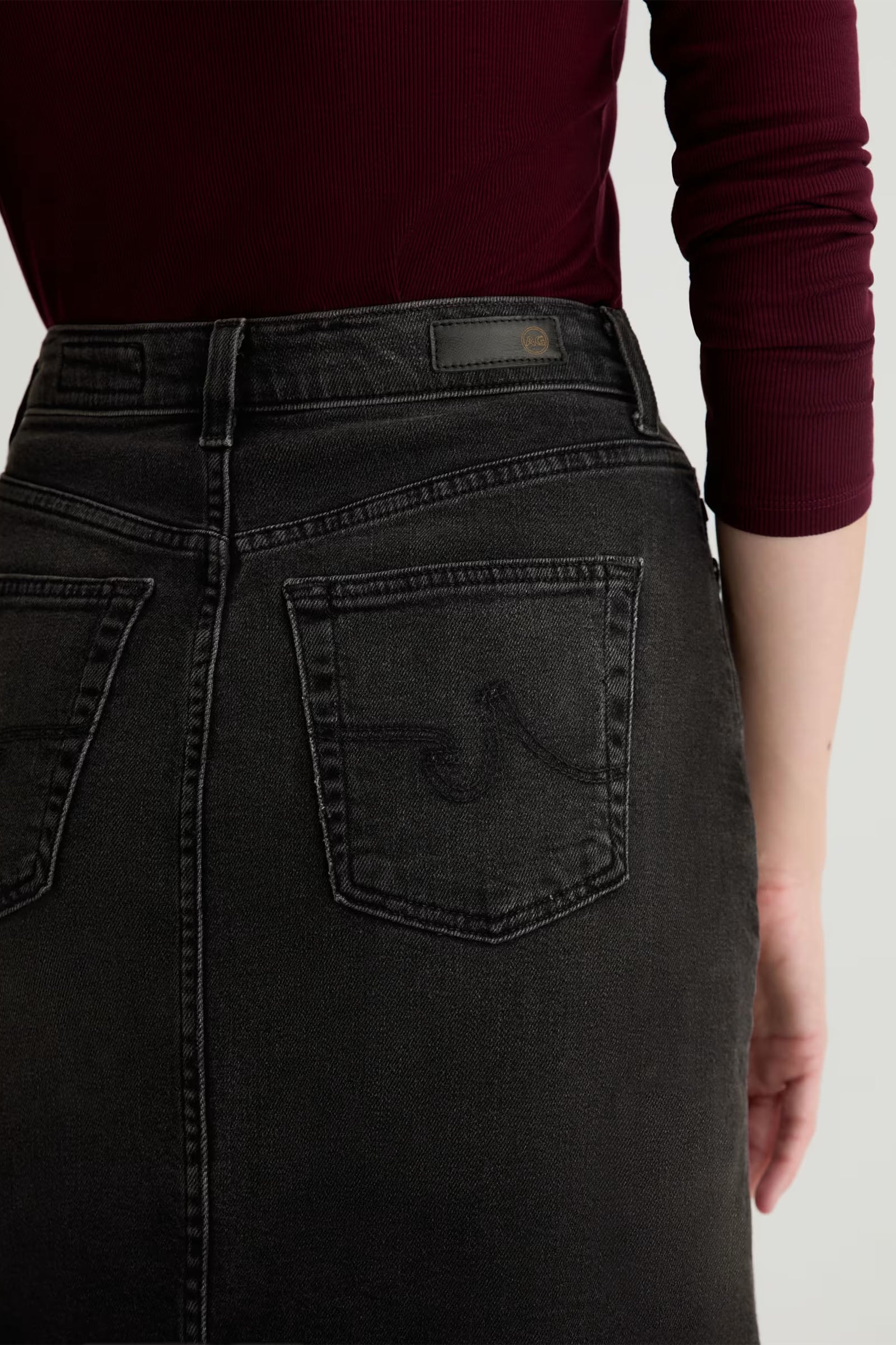 A person is shown from the back wearing a high-waisted Bruna Wrap Midi Skirt in black denim, highlighting the skirt’s pockets and detailed stitching, paired with a dark red long-sleeve top.