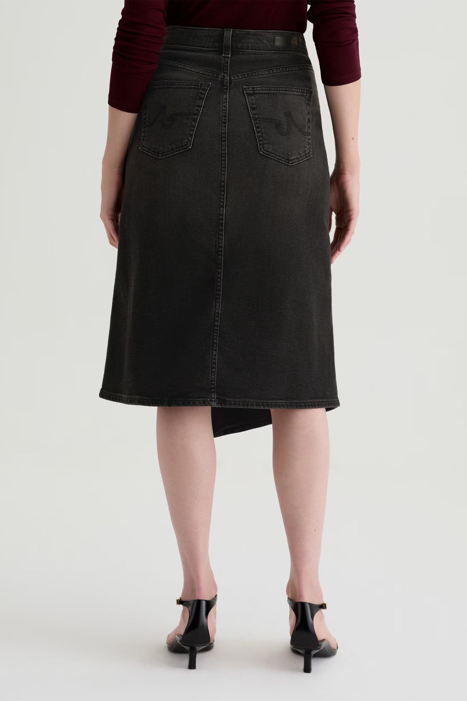A person is seen from behind wearing the Bruna Wrap Midi Skirt in black with back pockets, paired with a maroon top and black heeled sandals.
