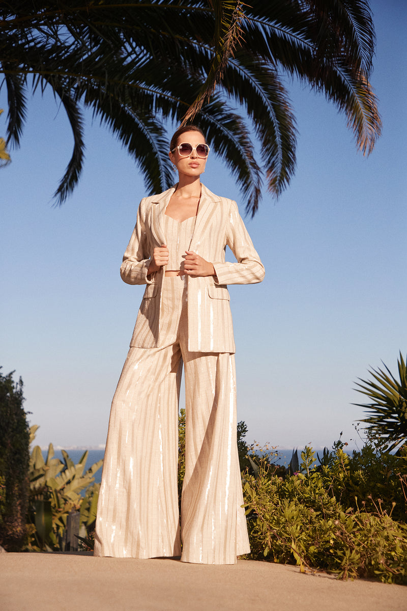 A woman stands confidently outdoors in the Alternates 2 Blazer, a stylish beige pinstripe suit, accessorized with sunglasses amid palm trees and a blue sky.
