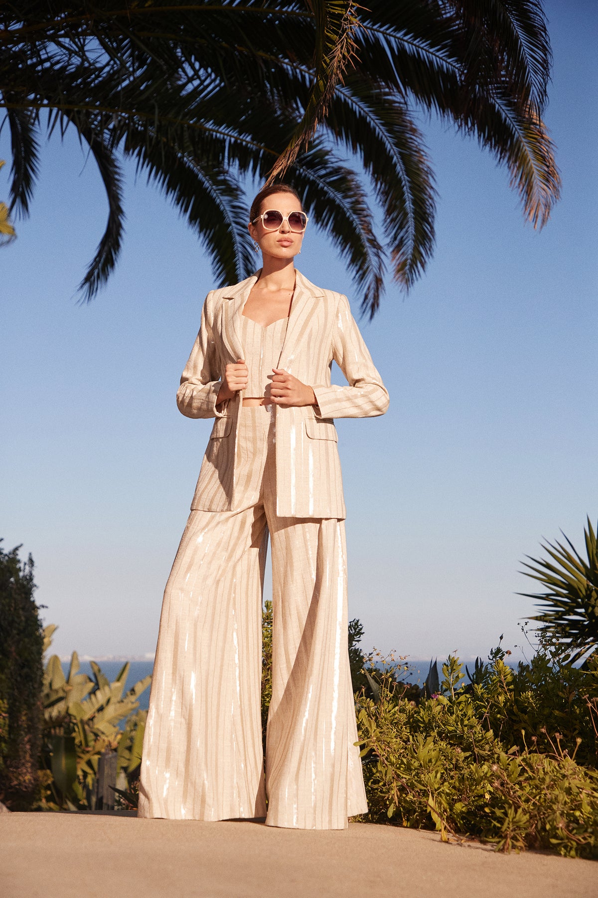 A woman stands outdoors under palm trees, wearing Nice Wide-Leg Pants as part of a beige pinstripe suit and sunglasses. She poses confidently, holding her jacket lapels with the ocean and clear blue sky in the background.