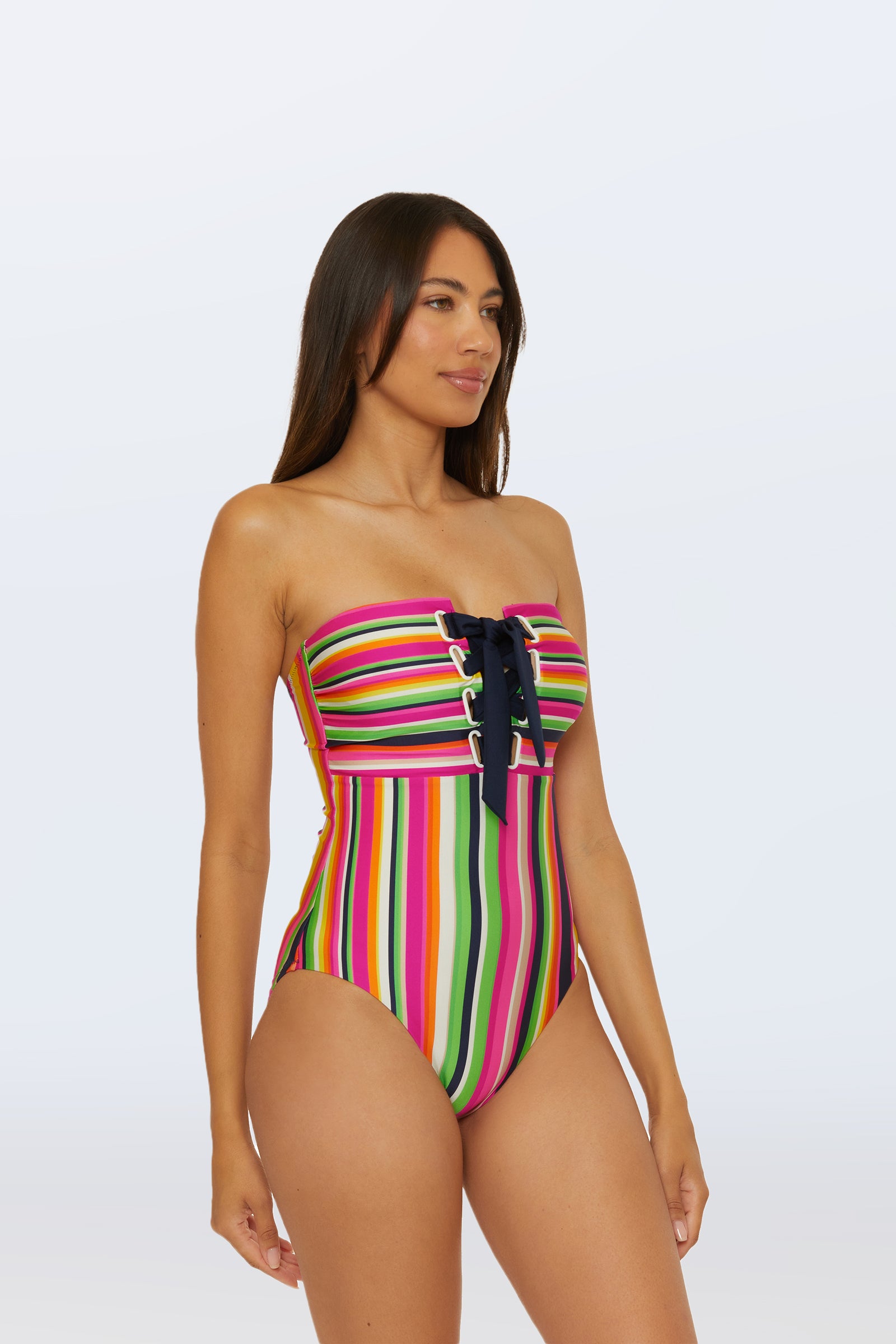 A woman models the Bandeau One-Piece Swimsuit - Splash Stripe, featuring multicolored stripes and a black lace-up front, while standing against a plain light background and looking slightly to the side.