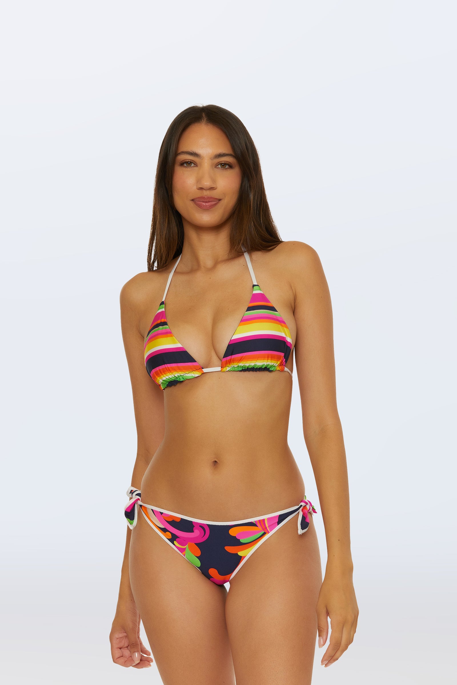 A woman with long brown hair wears the Reversible Tie Side Swim Bottom - Splash Stripe, featuring a floral pattern and side ties, paired with a striped bikini top, standing against a plain light background.