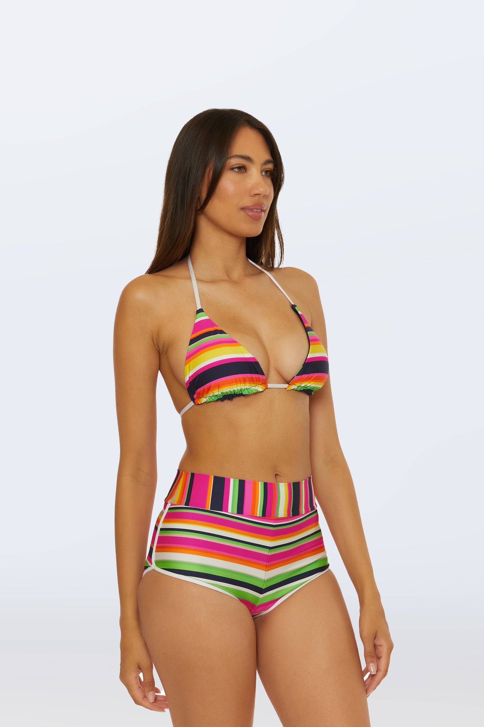 A woman with long brown hair wears the Boy Short Swim Bottom in Splash Stripe, featuring a colorful striped design, paired with a matching top and standing against a plain light background.
