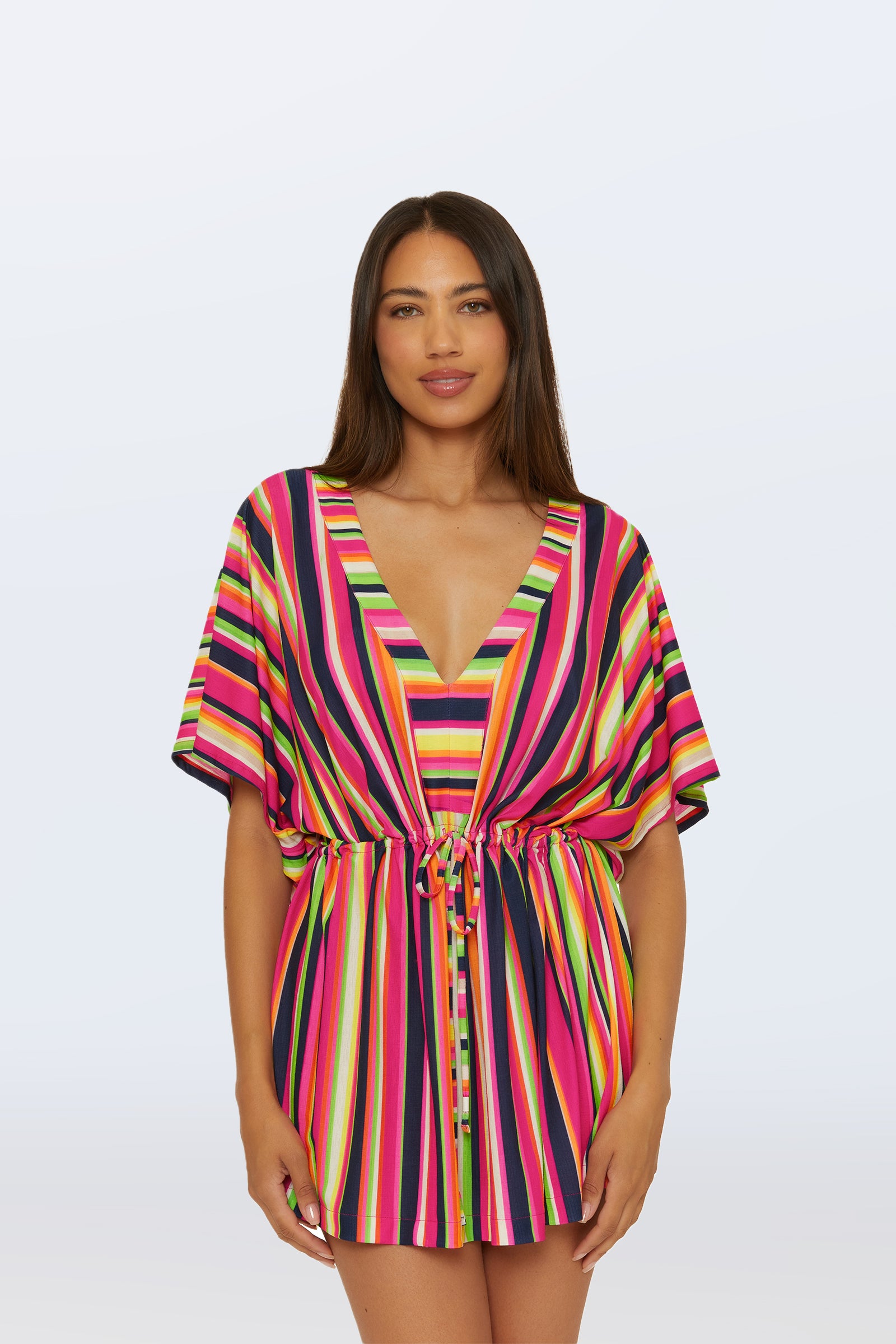 A woman faces forward in the Cinch Tunic V-Neck Cover-Up - Splash Stripe, a short, multi-colored dress with bright vertical stripes and a drawstring waist. The background is plain light gray.