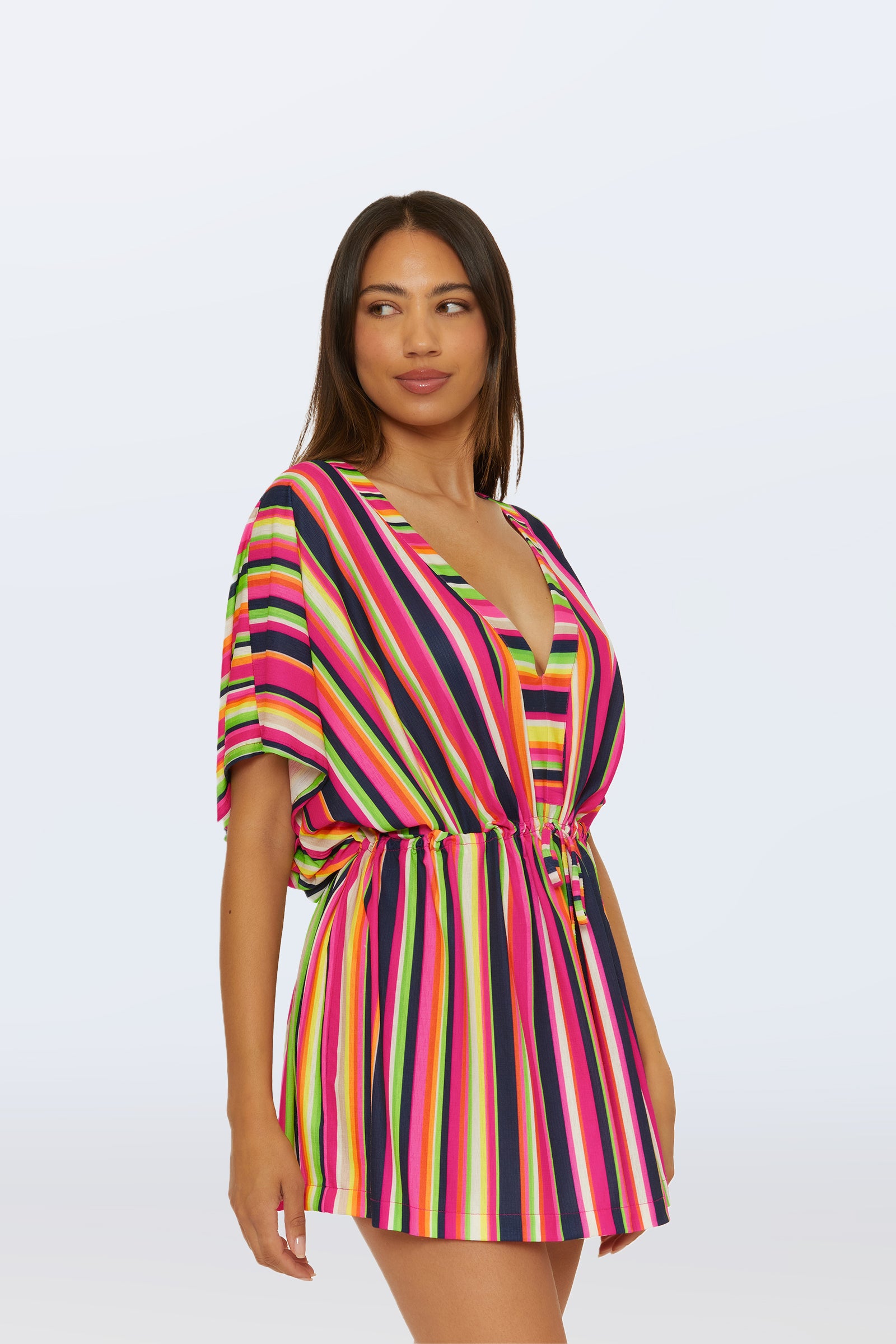 A woman poses against a light background wearing the Cinch Tunic V-Neck Cover-Up - Splash Stripe, a short, loose romper with vertical pink, yellow, green, and black stripes, a deep V-neck, and a cinched waist.