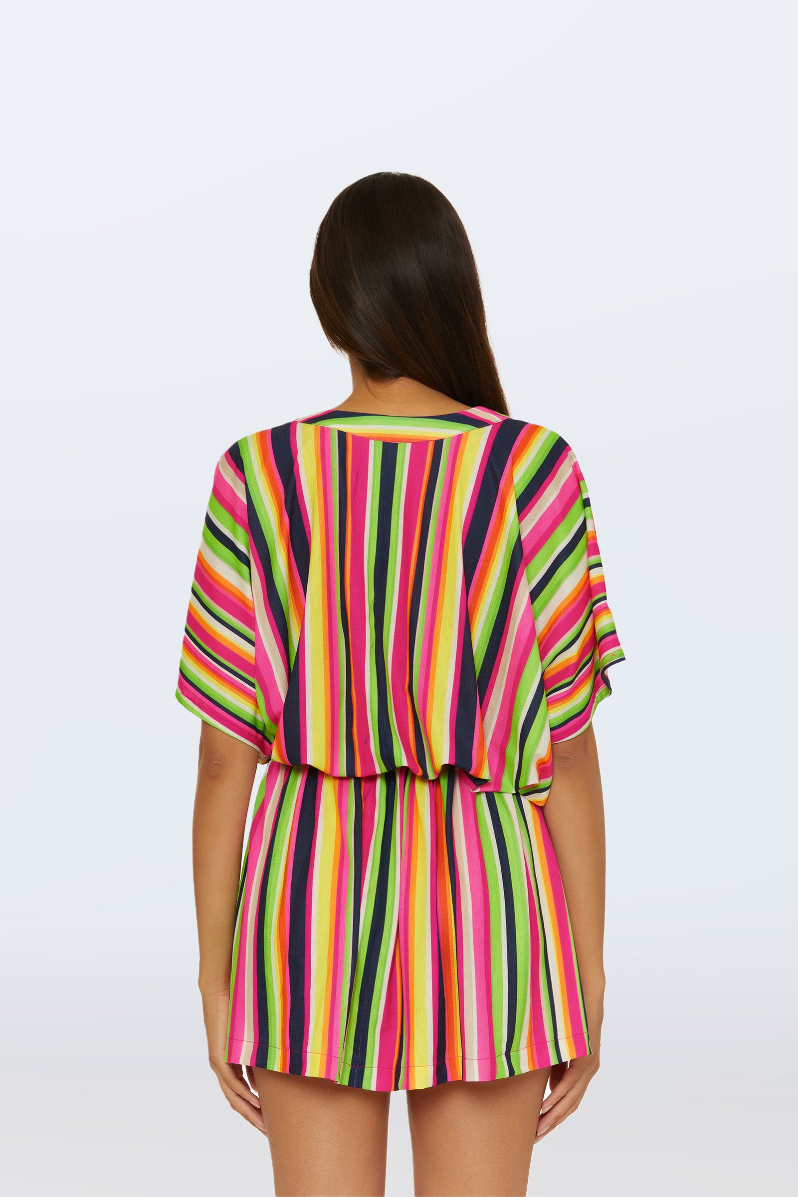 A woman with long, straight brown hair faces away from the camera, wearing the Cinch Tunic V-Neck Cover-Up in Splash Stripe—a colorful short-sleeve romper featuring vertical stripes in pink, yellow, green, purple, and white.
