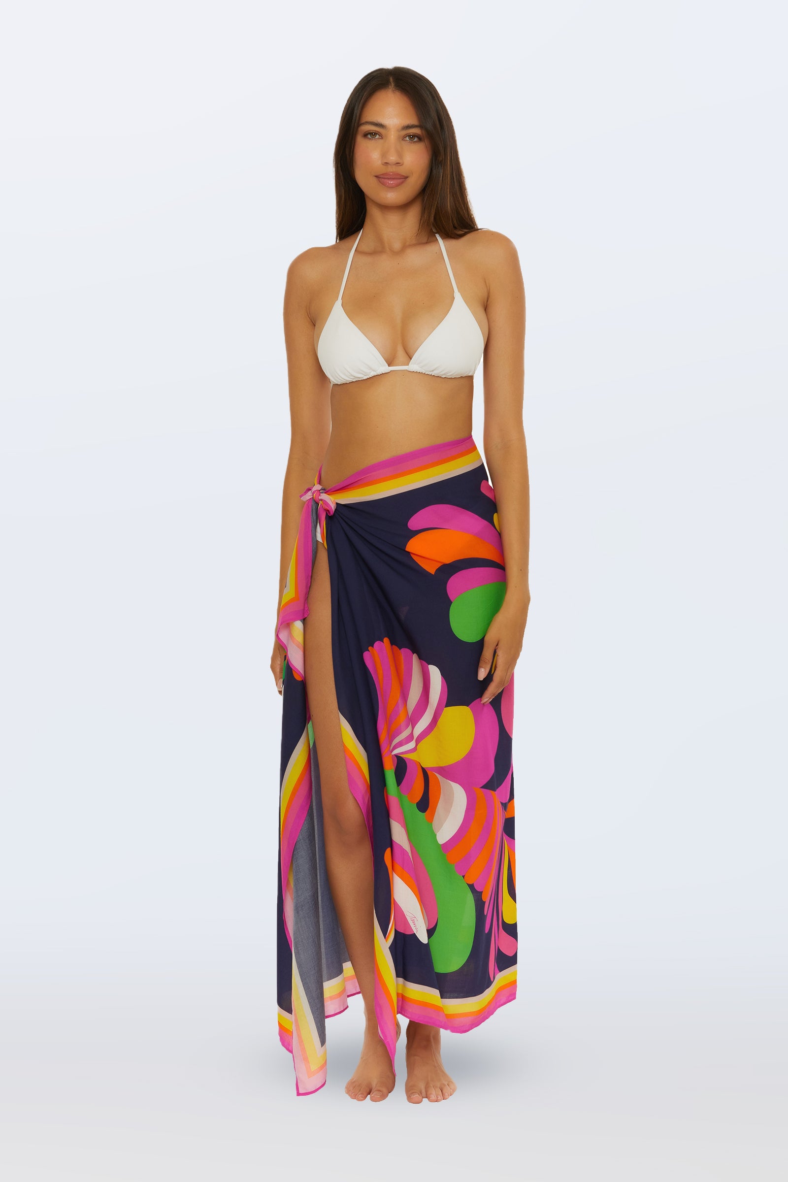 A woman faces forward wearing a white bikini top and the Pareo Swim Cover-Up - Palms Paradise, featuring vibrant pink, yellow, orange, and green palm-inspired patterns, set against a light background.