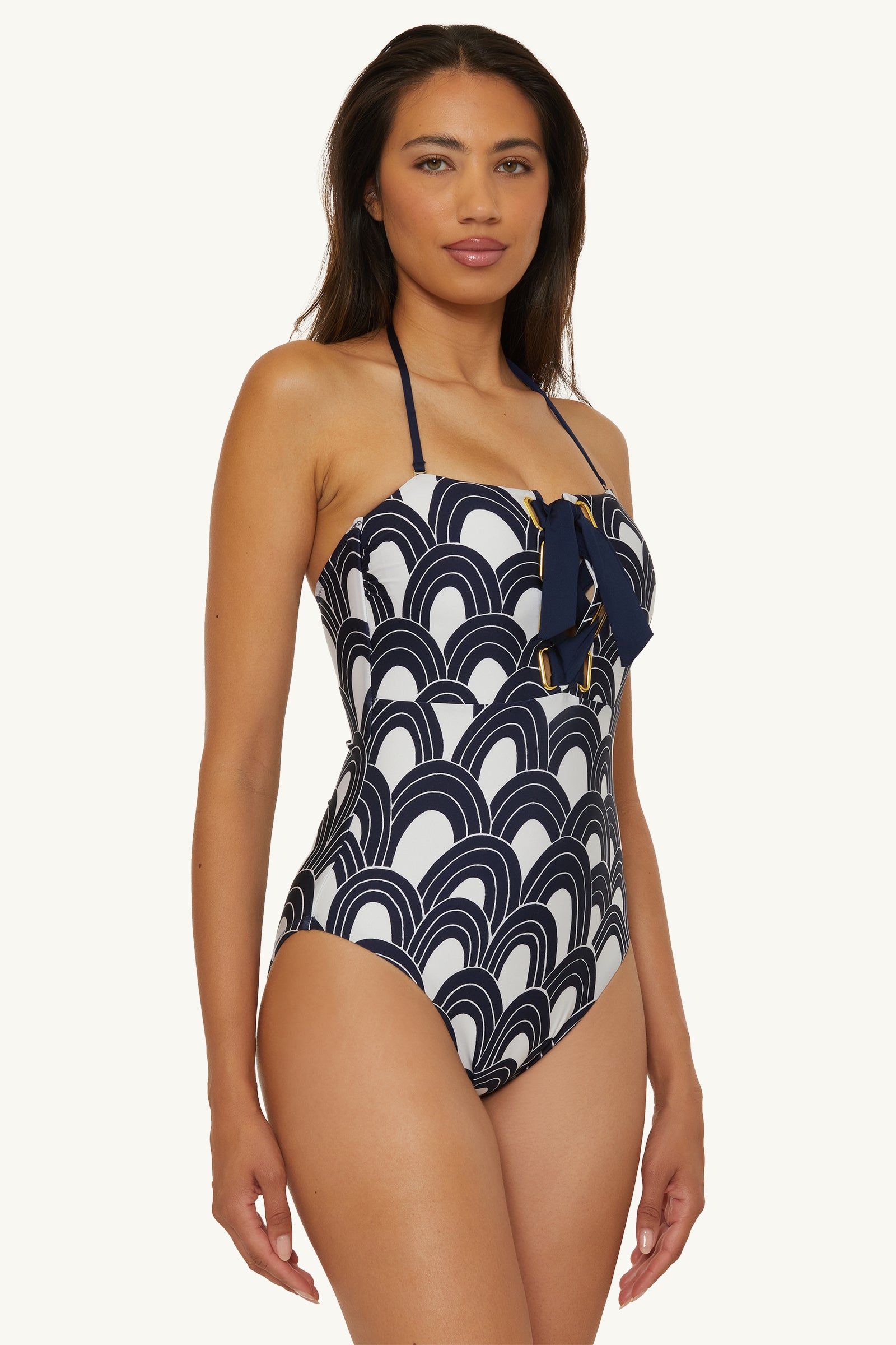 A woman wears the Lace-Up Bandeau One-Piece Swimsuit - Arches, featuring a navy and white geometric wave pattern, decorative bow on the chest, and halter-neck style, standing against a plain background.