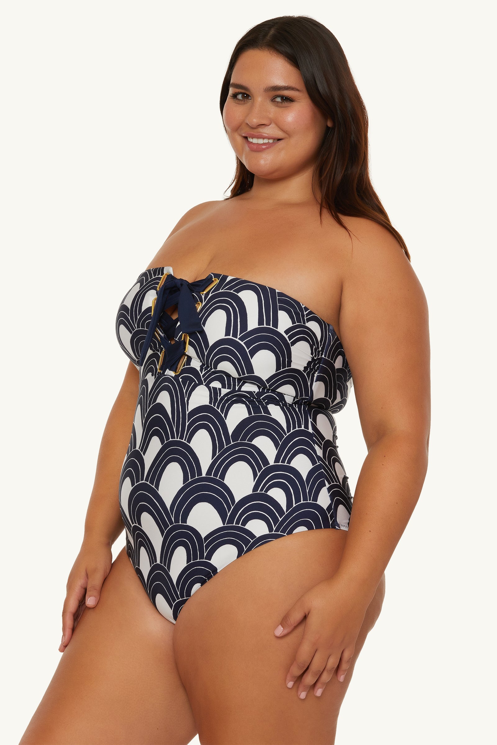 A smiling woman wears the Lace-Up Bandeau One-Piece Swimsuit - Arches, featuring a navy and white geometric wave pattern with a front tie detail, standing against a plain light background.