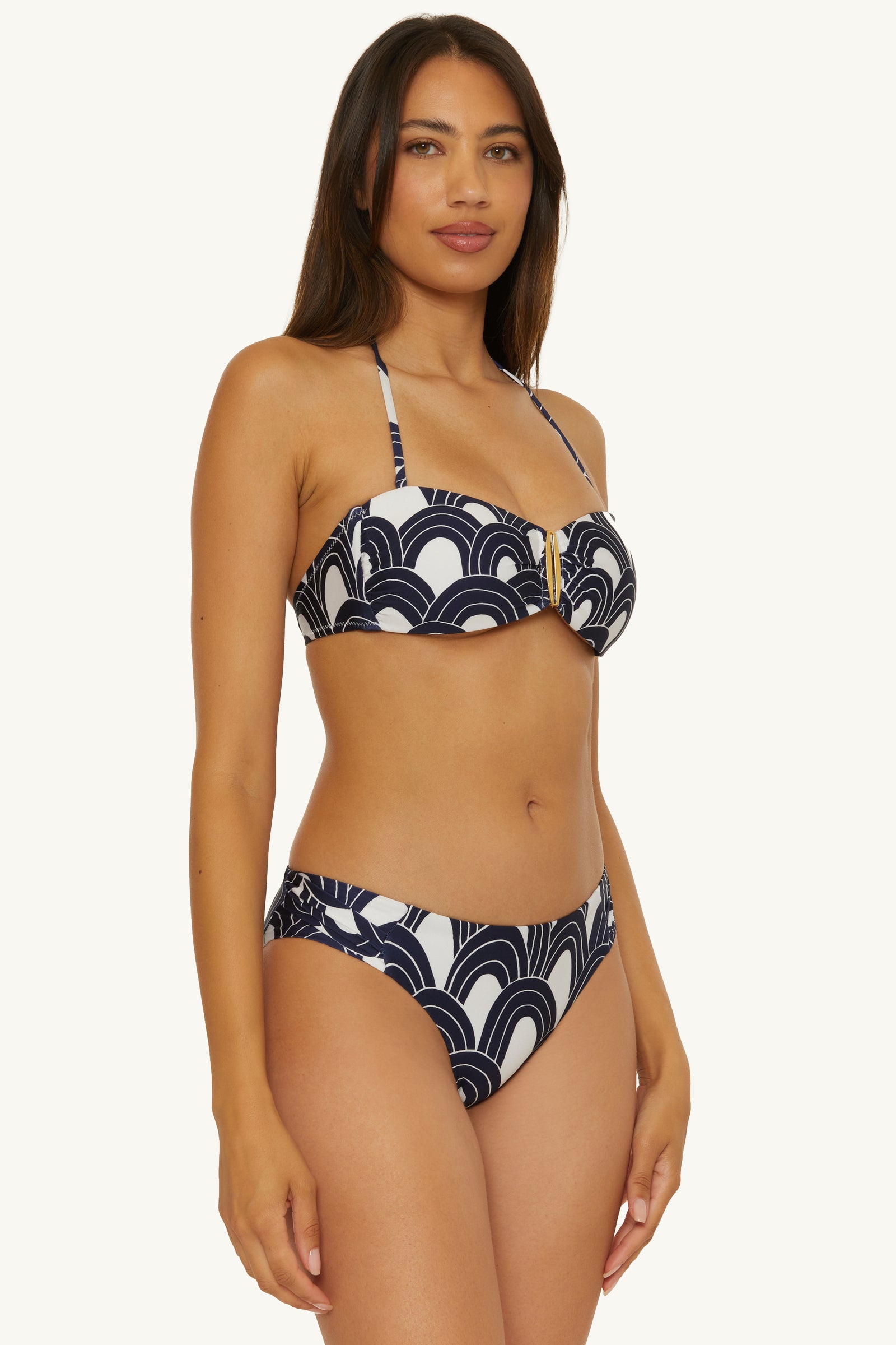 A woman wears the Buckle Bandeau Swim Top - Arches, featuring a black and white pattern with a gold center accent, standing against a plain light backdrop and looking at the camera.