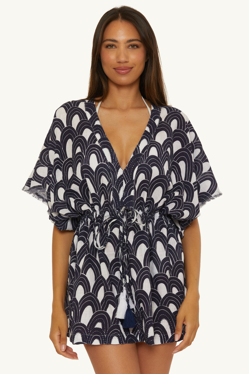 A woman with long, straight hair wears the V-Neck Cover-up Tunic Dress in Arches Swim, featuring a short length, tie waist, wide short sleeves, and a scallop pattern. She stands facing forward and smiles softly.