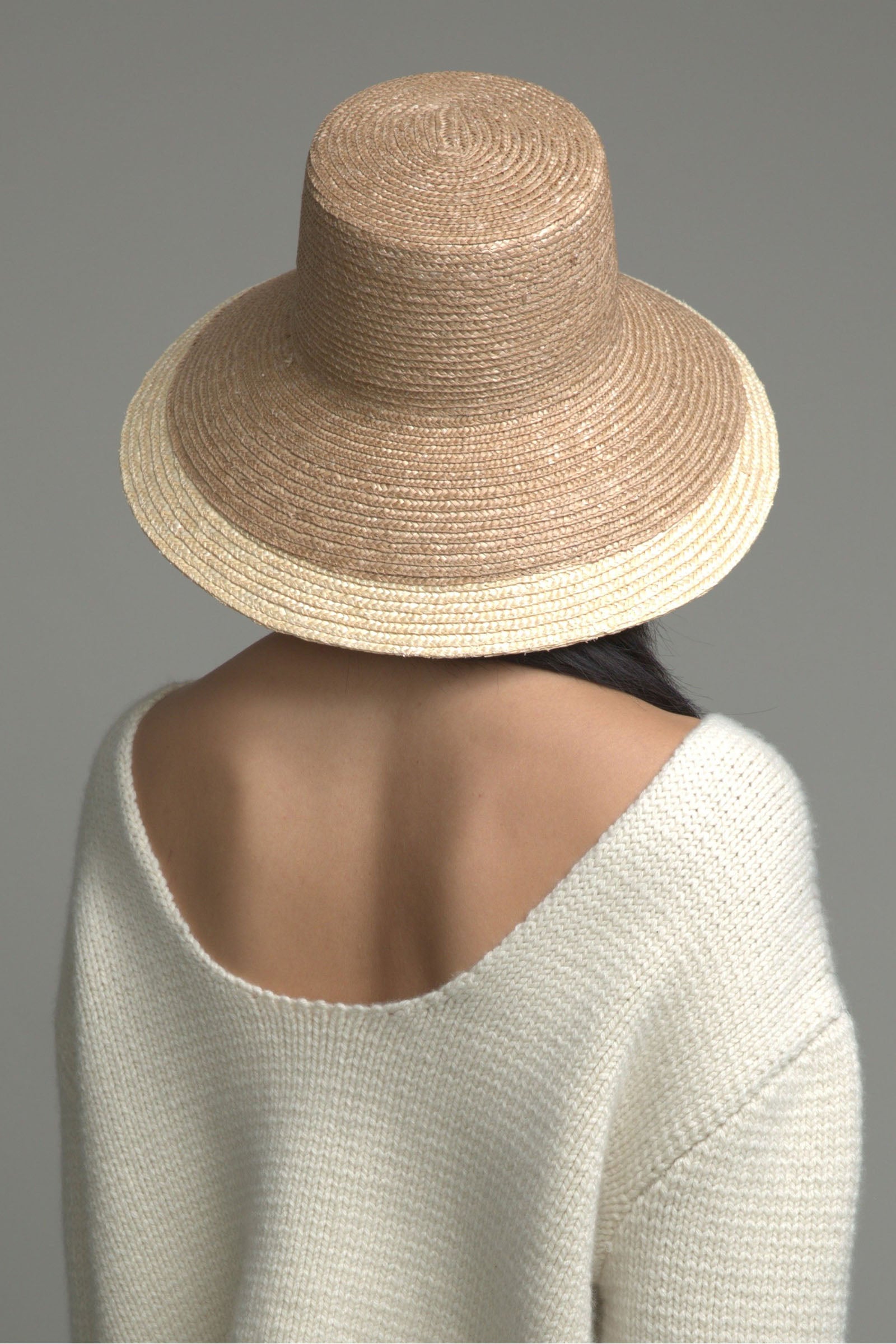 A person with long dark hair, seen from behind, wears the Eugenia Kim Annabelle Hat and an off-the-shoulder cream-colored knitted sweater against a plain gray background.