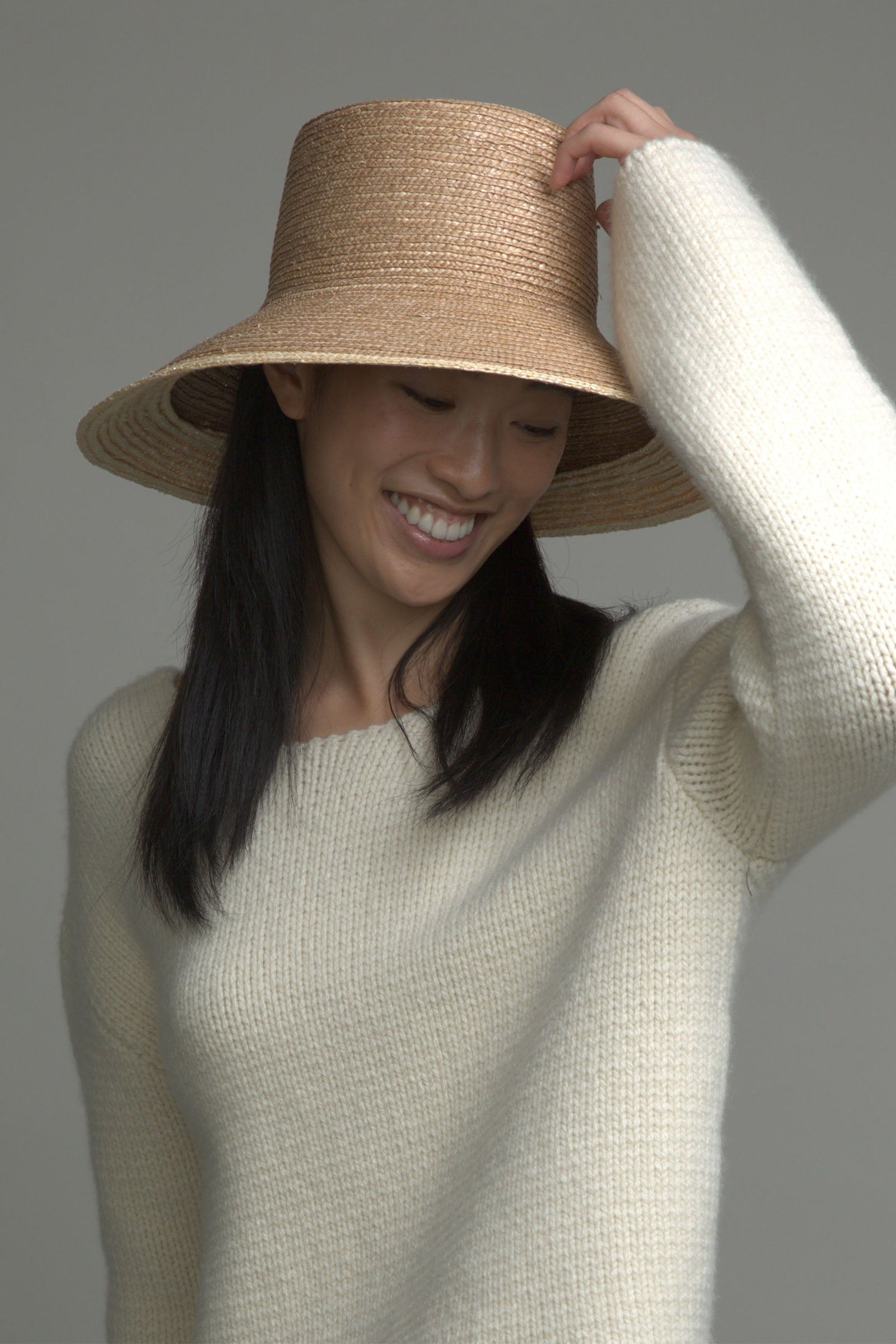 A woman with long dark hair smiles while looking down, wearing the Eugenia Kim Annabelle Hat—a wide-brimmed straw sun hat with UPF 50+ protection—and a cream knit sweater, raising her left hand to the brim against a gray background.