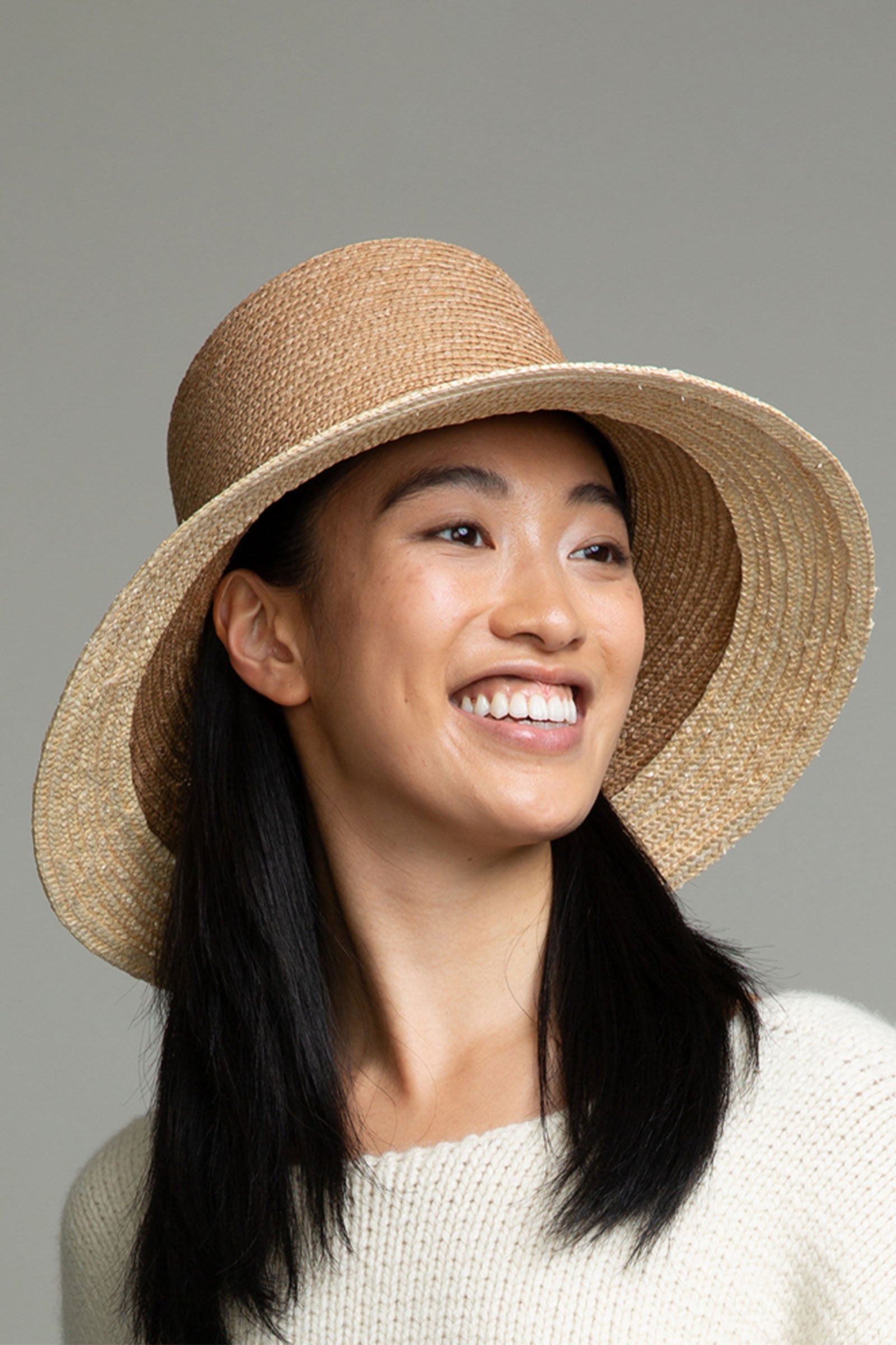 A smiling person with long black hair wears the Eugenia Kim Annabelle Hat and a light-colored sweater against a plain gray background.