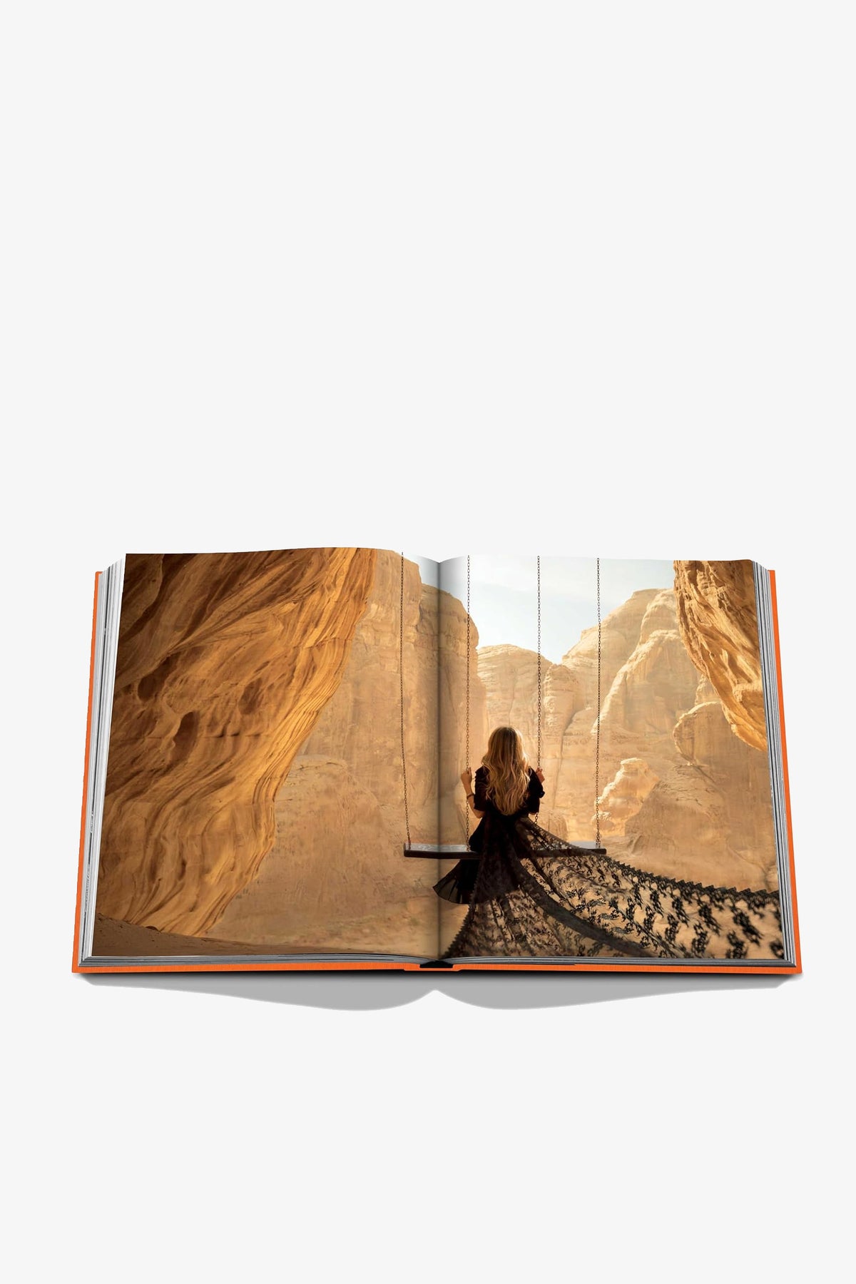 An open Assouline Alula Ever Book displaying a photo of a woman in a long, dark dress sitting on a swing, surrounded by tall sandstone cliffs under a clear sky.