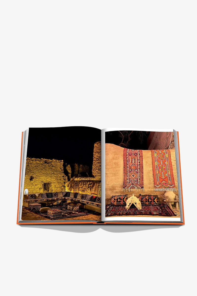 An open Assouline Alula Ever Book displaying photographs of a rustic room with woven rugs, stone walls, and pottery, set against a plain white background.