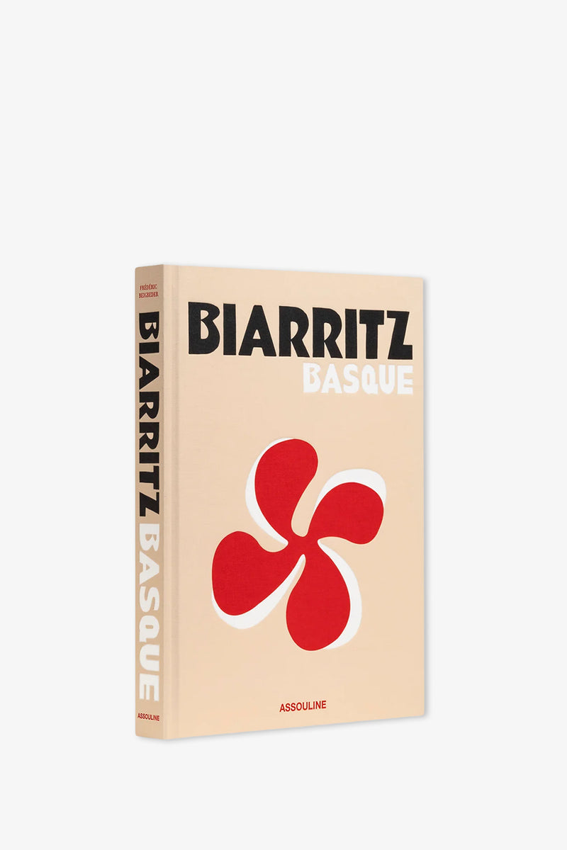 The Assouline Biarritz Basque Book is a beige hardcover featuring a striking red Basque cross symbol on its cover. The book’s spine displays the title, while the publisher, ASSOULINE, is printed at the bottom.
