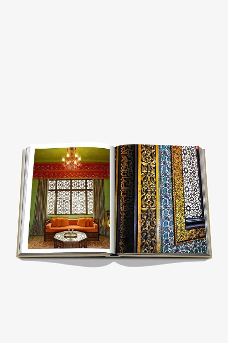 An open Assouline Cairo Eternal book displays an ornate interior with an orange sofa, carved wooden window screens and a chandelier on the left page, and richly patterned gold and blue textiles on the right.