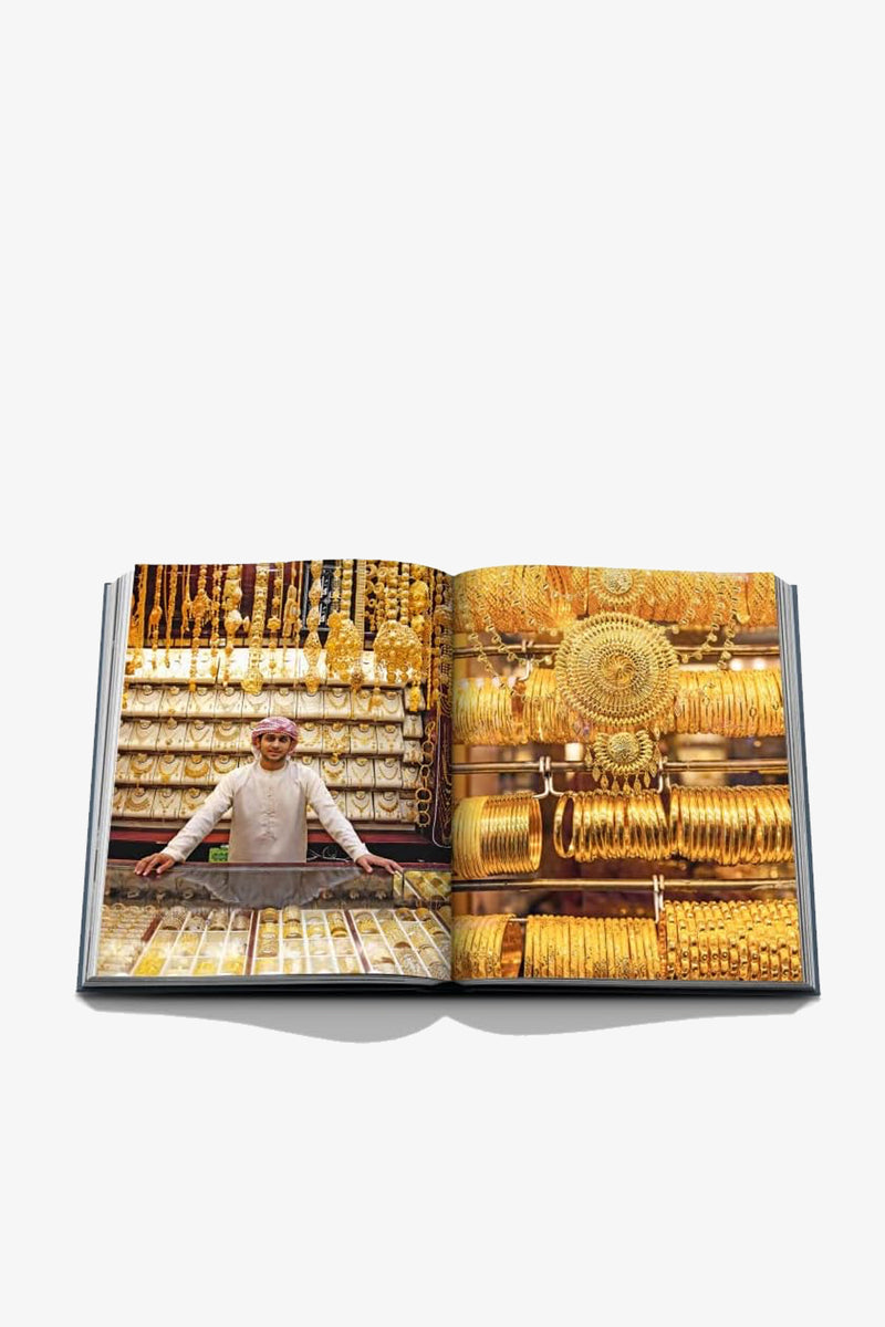 An open copy of “Assouline Dubai Wonder,” featuring an image of a man standing behind a counter filled with gold jewelry, with shelves behind him adorned with numerous gold necklaces, bangles, and ornaments.