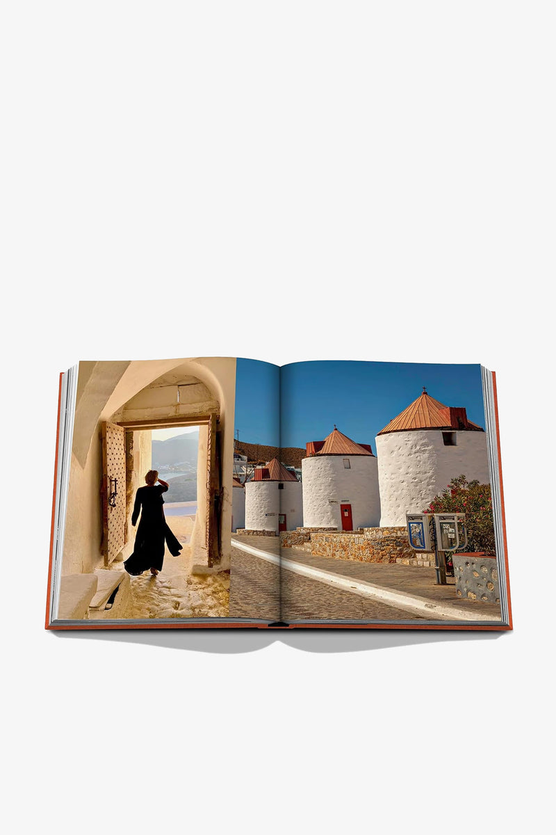 The open pages of ASSOULINE GREEK ISLANDS reveal two images: one on the left of a person walking beneath an archway toward the sea, and another on the right showcasing traditional white windmills with red roofs set against a vivid blue sky.