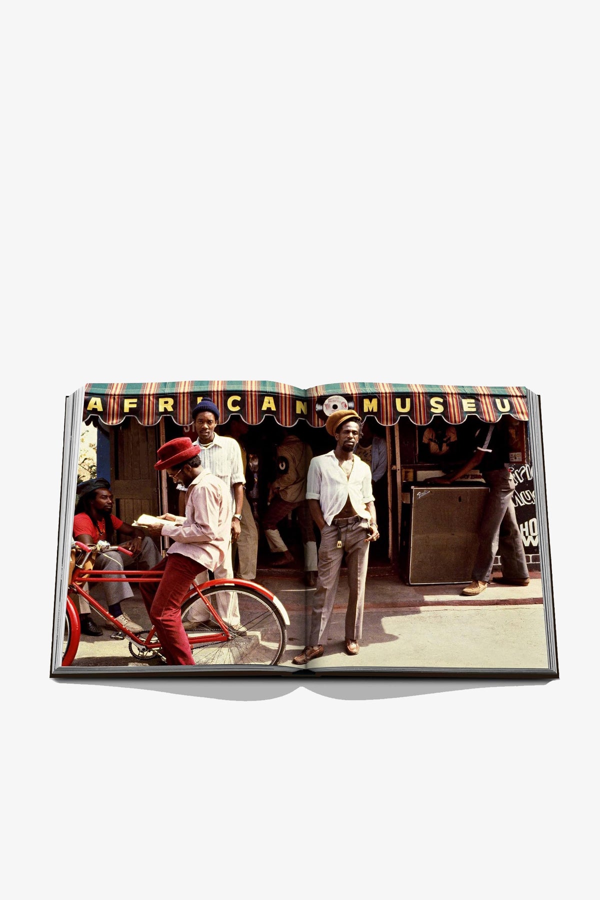 An open copy of Assouline Jamaica Vibes displays a photo of people standing and sitting outside a building with a striped awning that reads AFRICAN MUSEUM; one person is on a red bicycle.