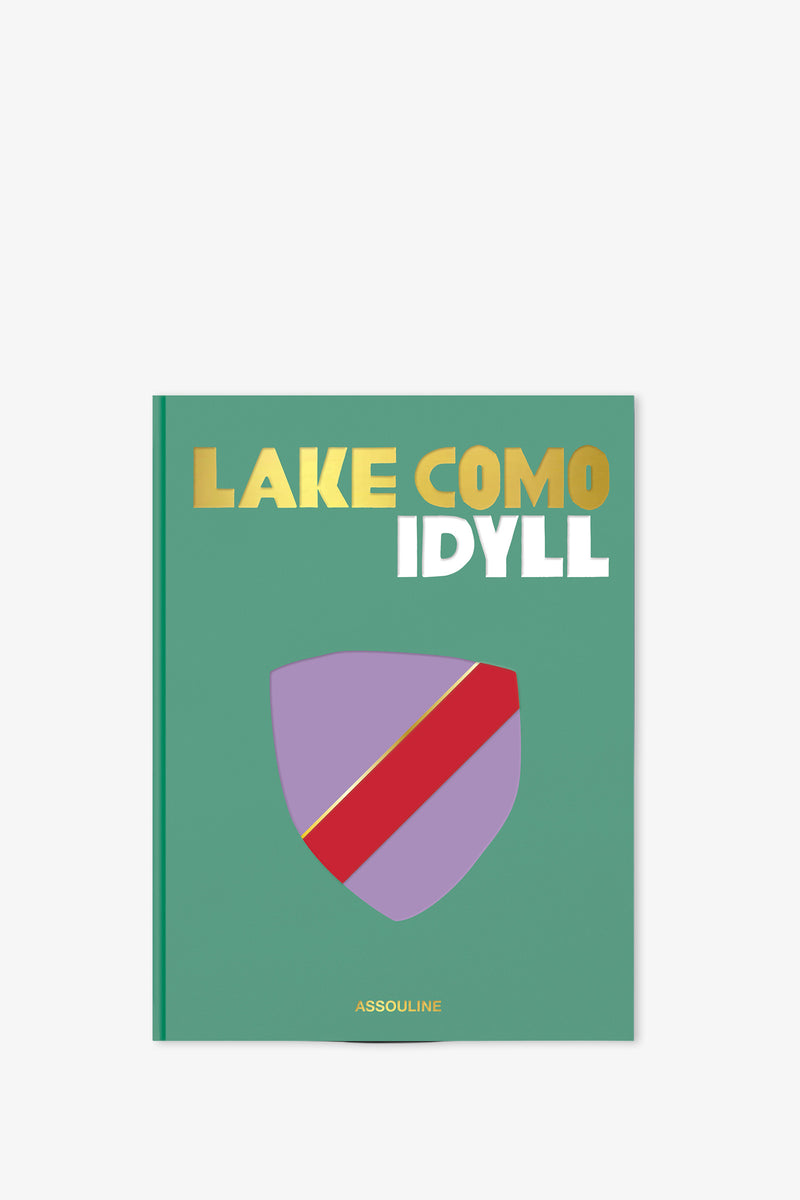 The Assouline Lake Como Idyll Book features a green cover with gold and white text, adorned with a purple shield bearing a red diagonal stripe and the publisher's name, Assouline, at the bottom.