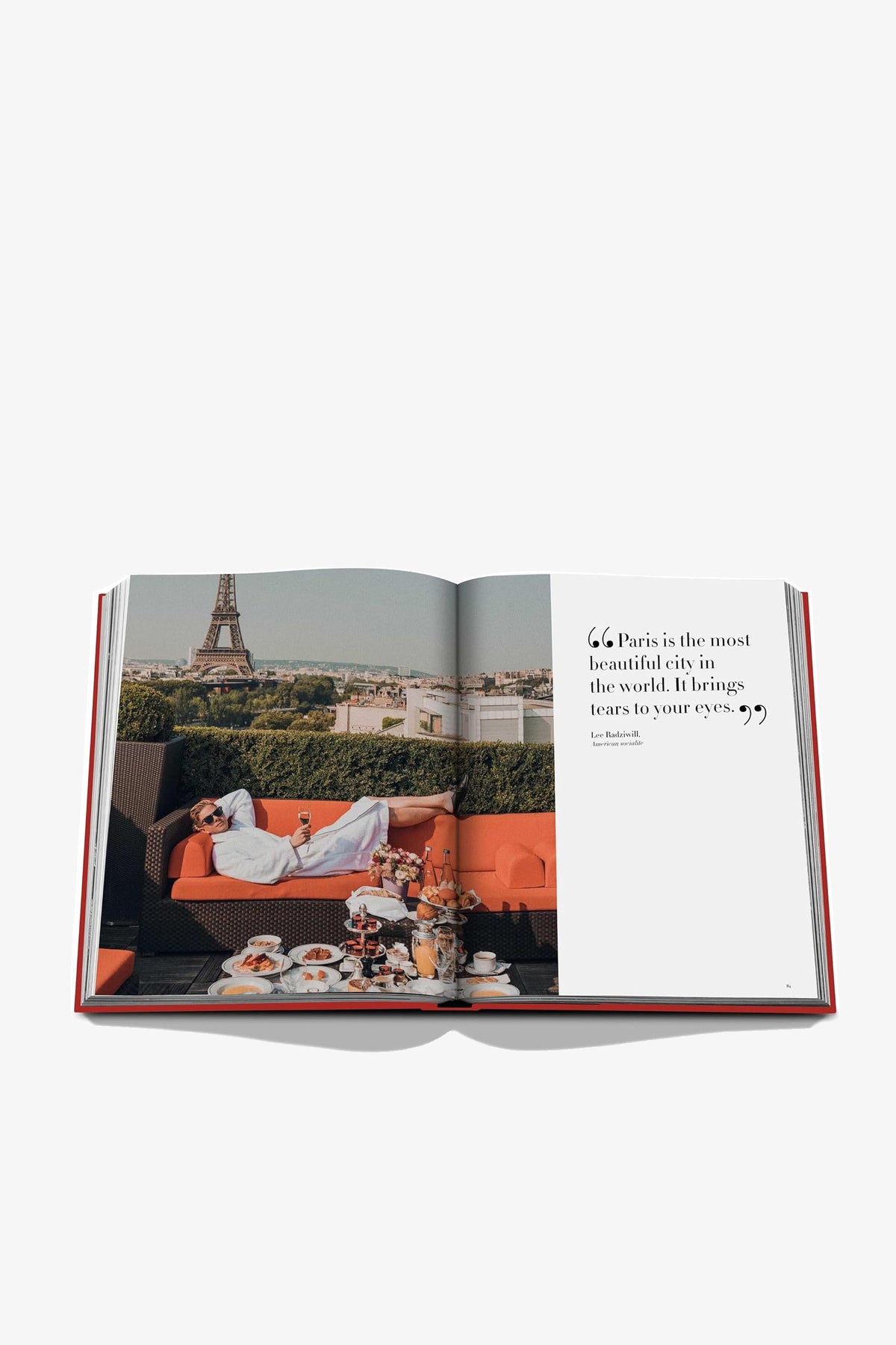 An open Assouline Plaza Athénée book displays a woman lounging on an orange sofa on a terrace with breakfast and a view of the Eiffel Tower on the left page, and a quote on the right page.