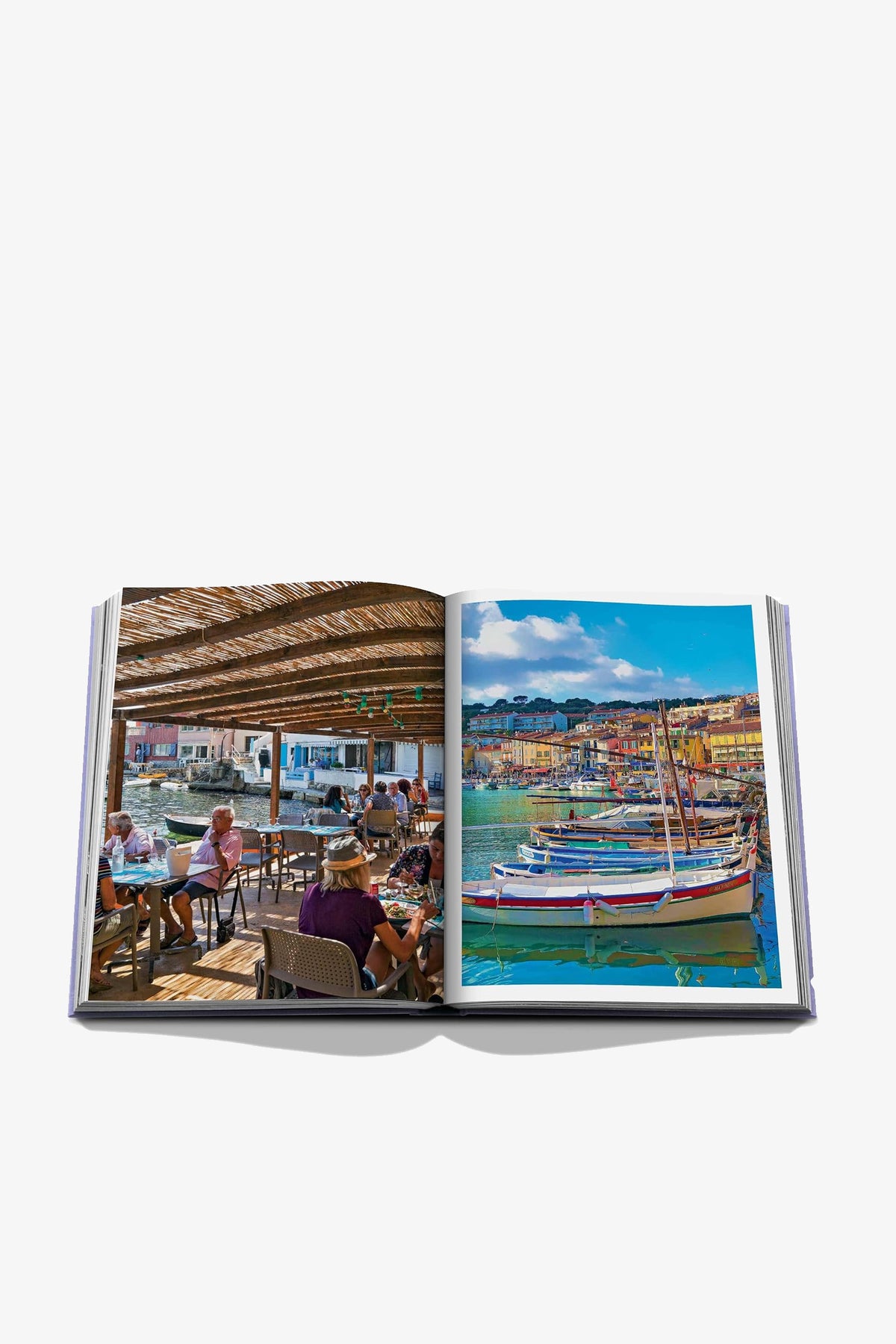 An open Assouline Provence Glory Book displays a photo of people dining outdoors by the water on the left page, and colorful boats docked at a vibrant harbor on the right page.