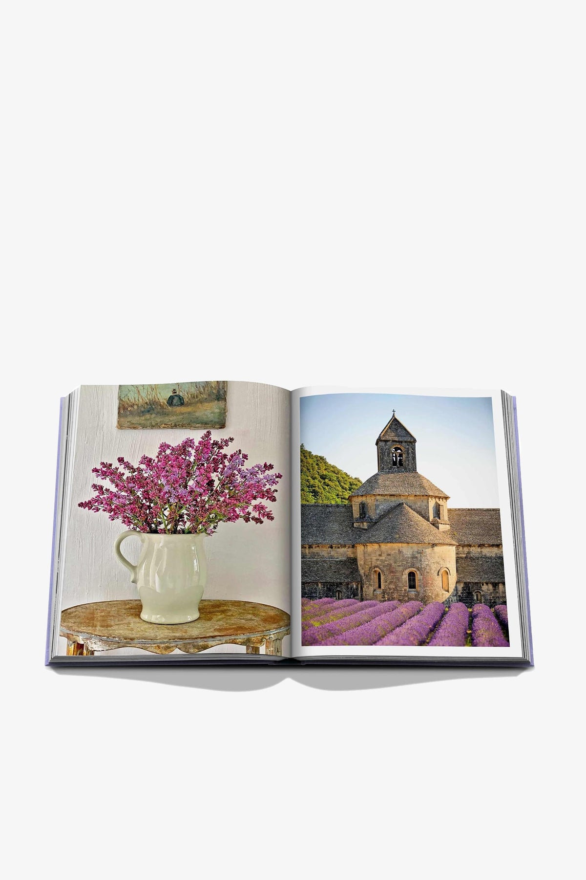 The Assouline Provence Glory Book lies open, revealing a photo of a vase with purple flowers on the left page and an image of a stone church set before a lavender field on the right.