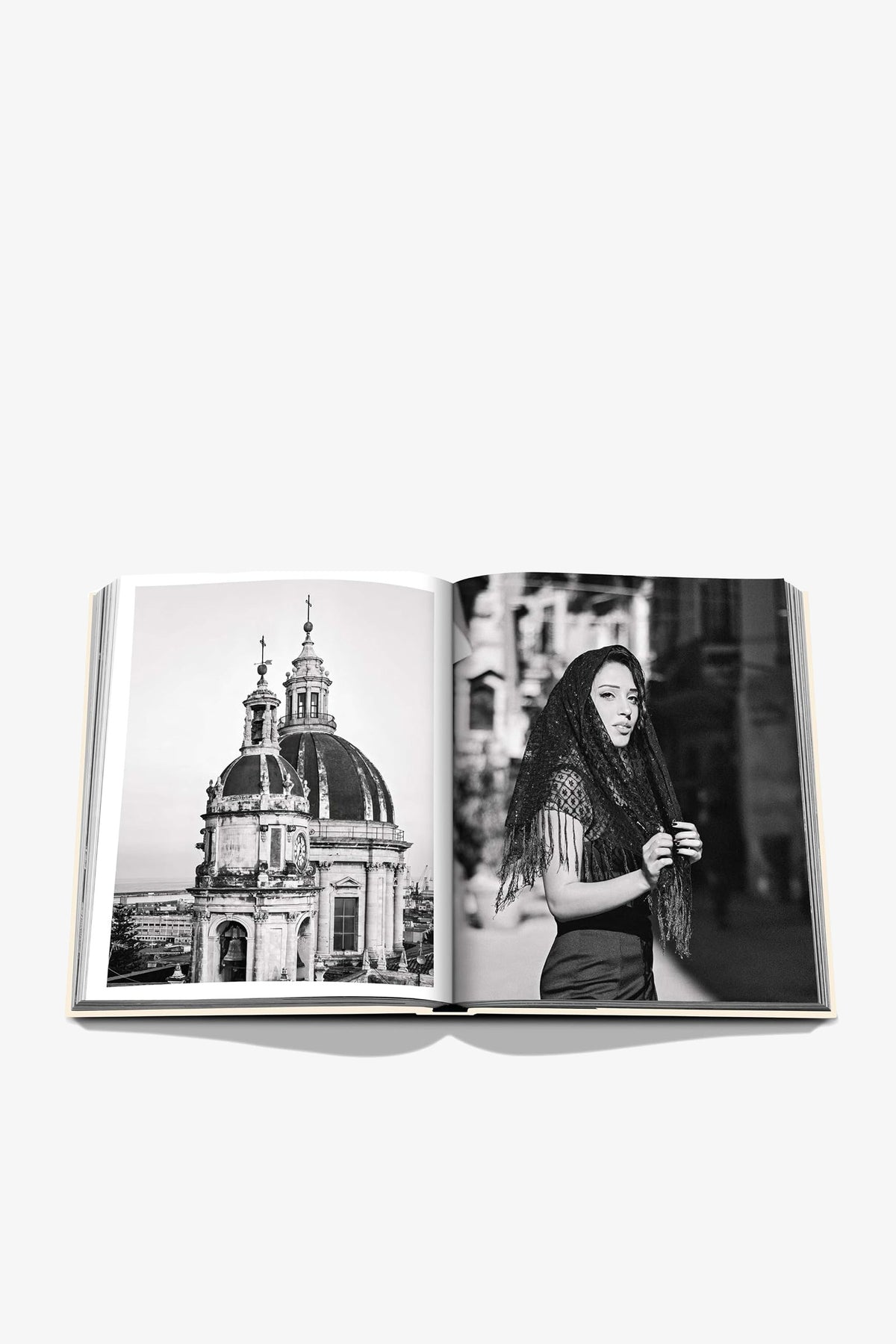 An open Assouline Sicily Honor book displays black-and-white photos of a domed Baroque cathedral on the left page and a woman with long dark hair and a fringed shawl on a city street on the right.