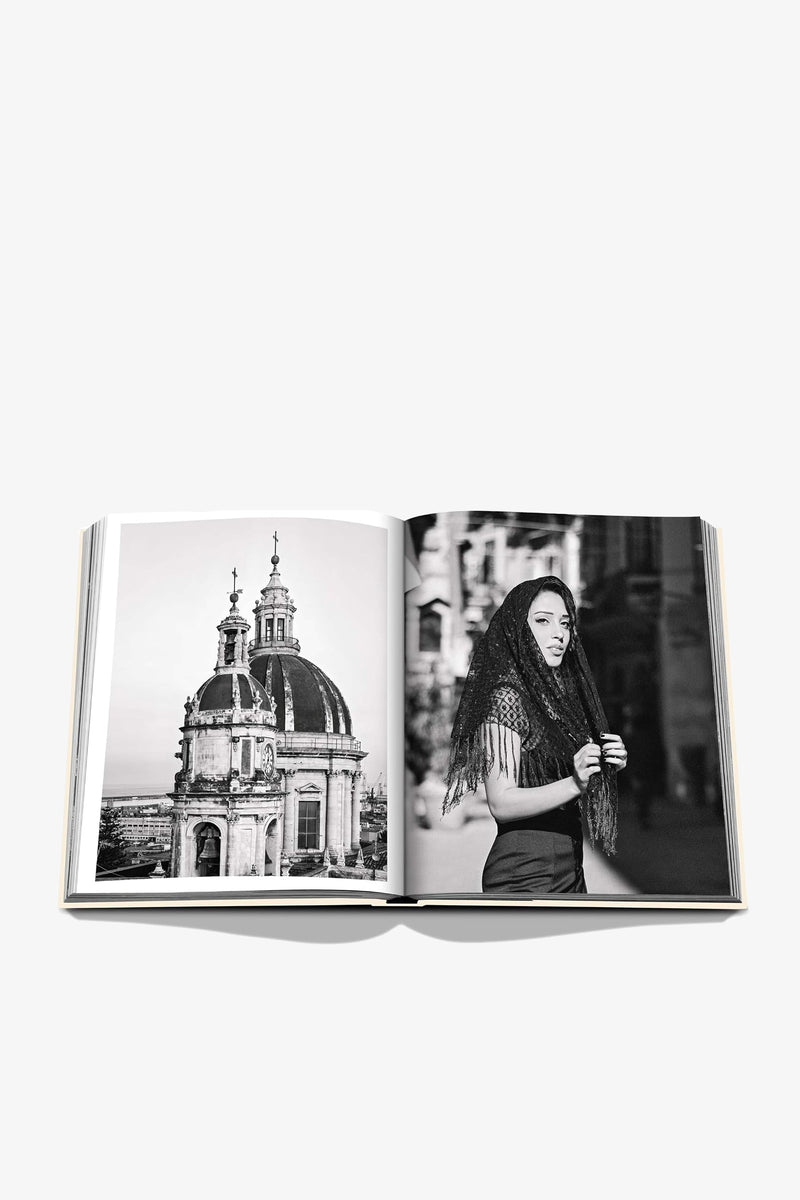 An open Assouline Sicily Honor book displays black-and-white photos of a domed Baroque cathedral on the left page and a woman with long dark hair and a fringed shawl on a city street on the right.