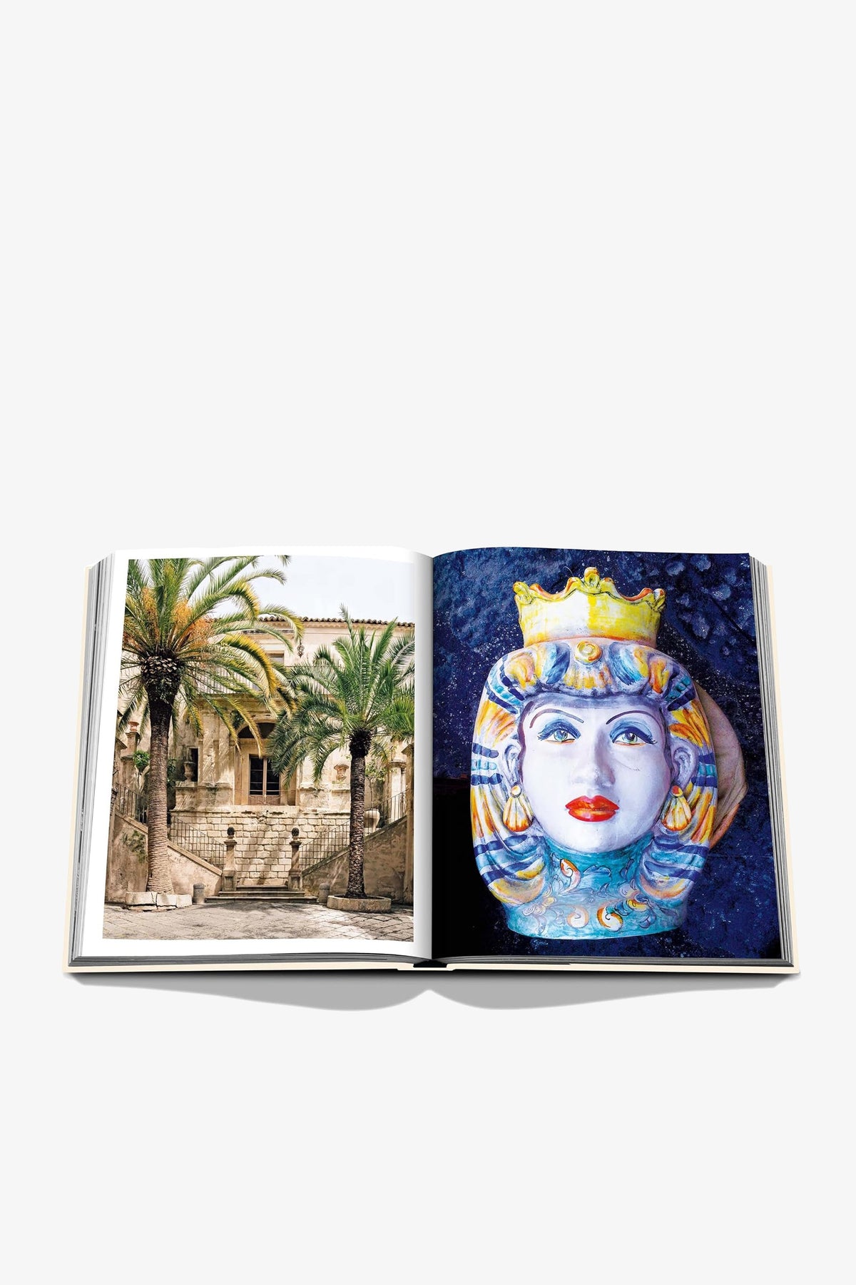 An open Assouline Sicily Honor book displays a sunlit stone villa framed by palm trees on the left page and a hand-painted Sicilian ceramic head vase with a gold crown on a deep blue background on the right.