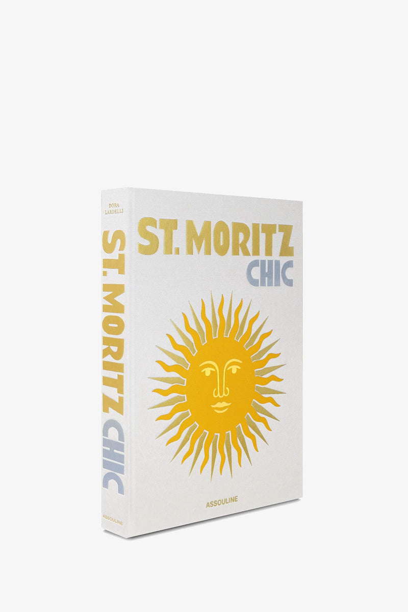 A white Assouline St Moritz Chic book shown at an angle, with the golden sun illustration on the cover and the title printed in gold and blue along the spine.