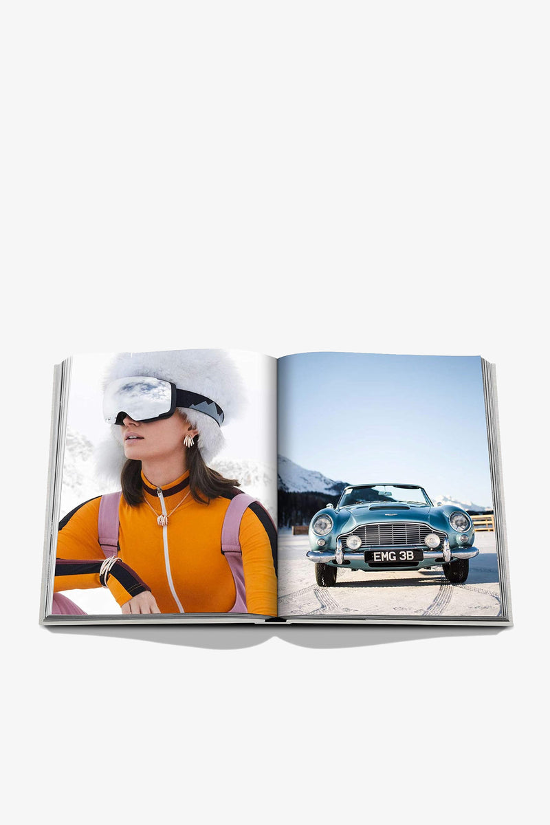 An open Assouline St Moritz Chic book displays a woman in an orange ski suit, white goggles and fur hat on the left page, and a vintage green convertible parked on a snowy road on the right.