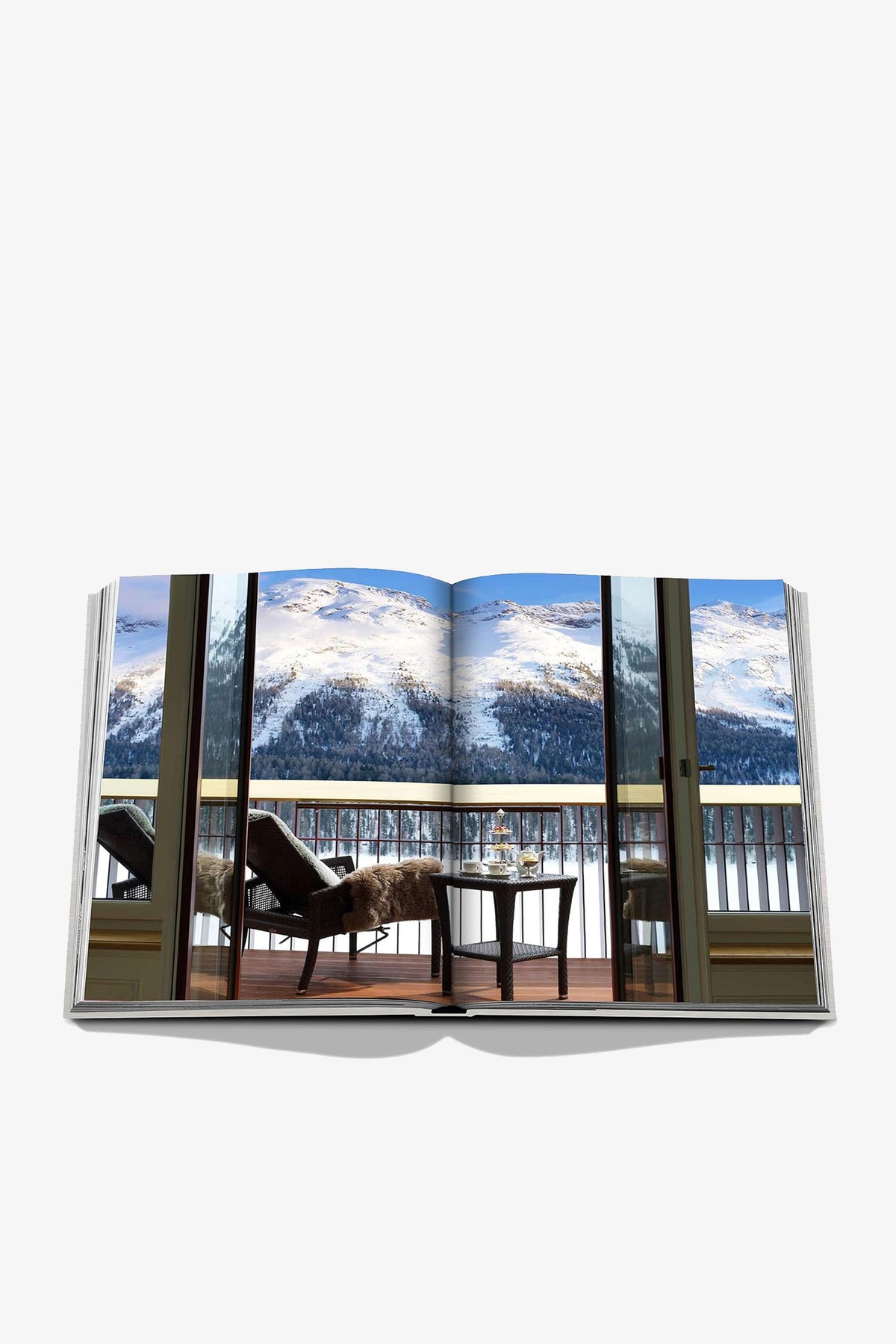 An open Assouline St Moritz Chic book displays a two-page photo of a balcony with lounge chairs and fur throws overlooking snow-capped alpine mountains.