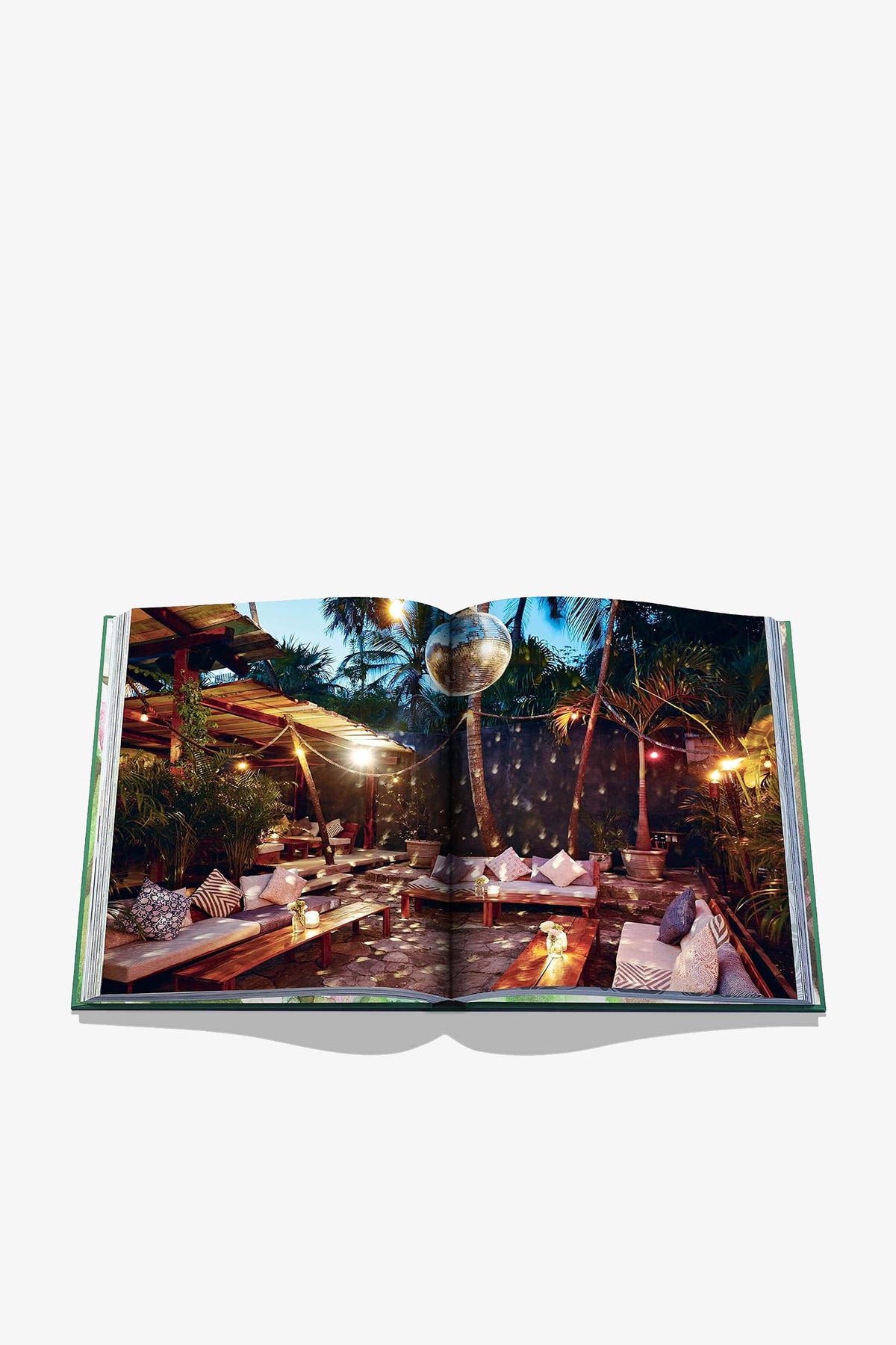 An open Assouline Tulum Gypset Book displays a two-page photograph of an outdoor lounge area at dusk, featuring low wooden seating, string lights, and a disco ball hanging in palm trees.