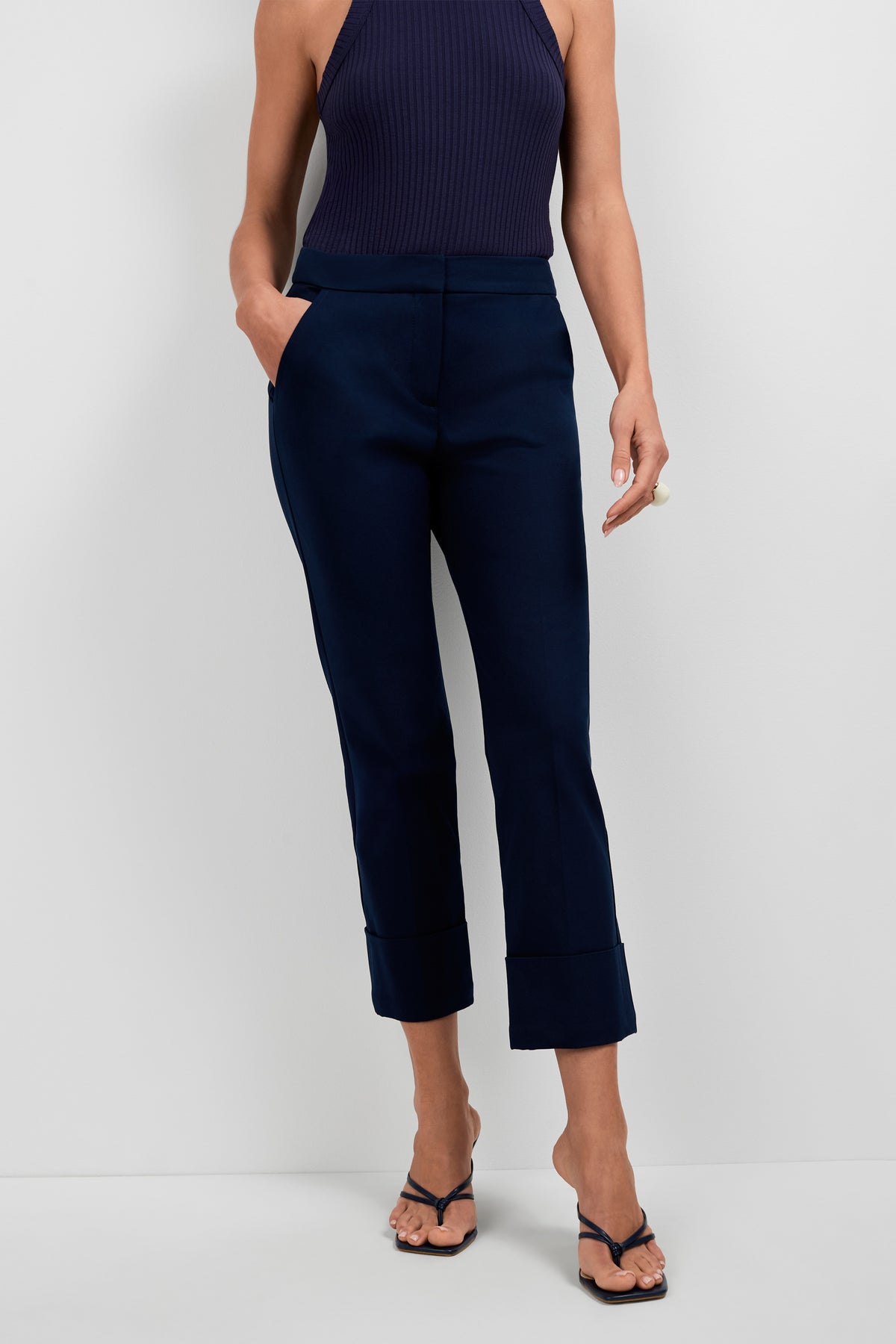 A woman stands against a plain white background, hands in her pockets, wearing Banshee Cropped Cuff Hem Pants in navy, a matching sleeveless top, and black heeled sandals.