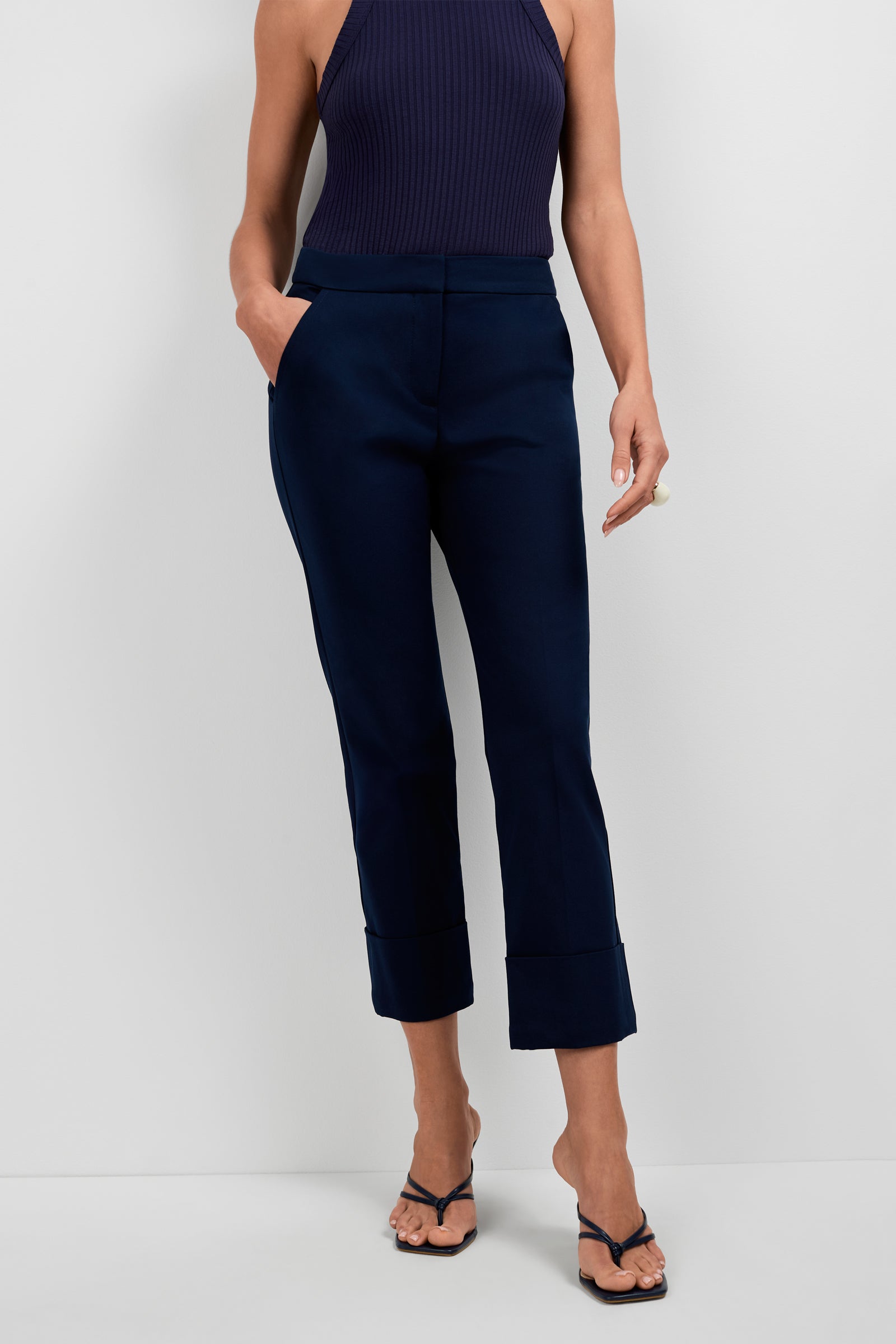 A woman stands against a plain white background, hands in her pockets, wearing Banshee Cropped Cuff Hem Pants in navy, a matching sleeveless top, and black heeled sandals.