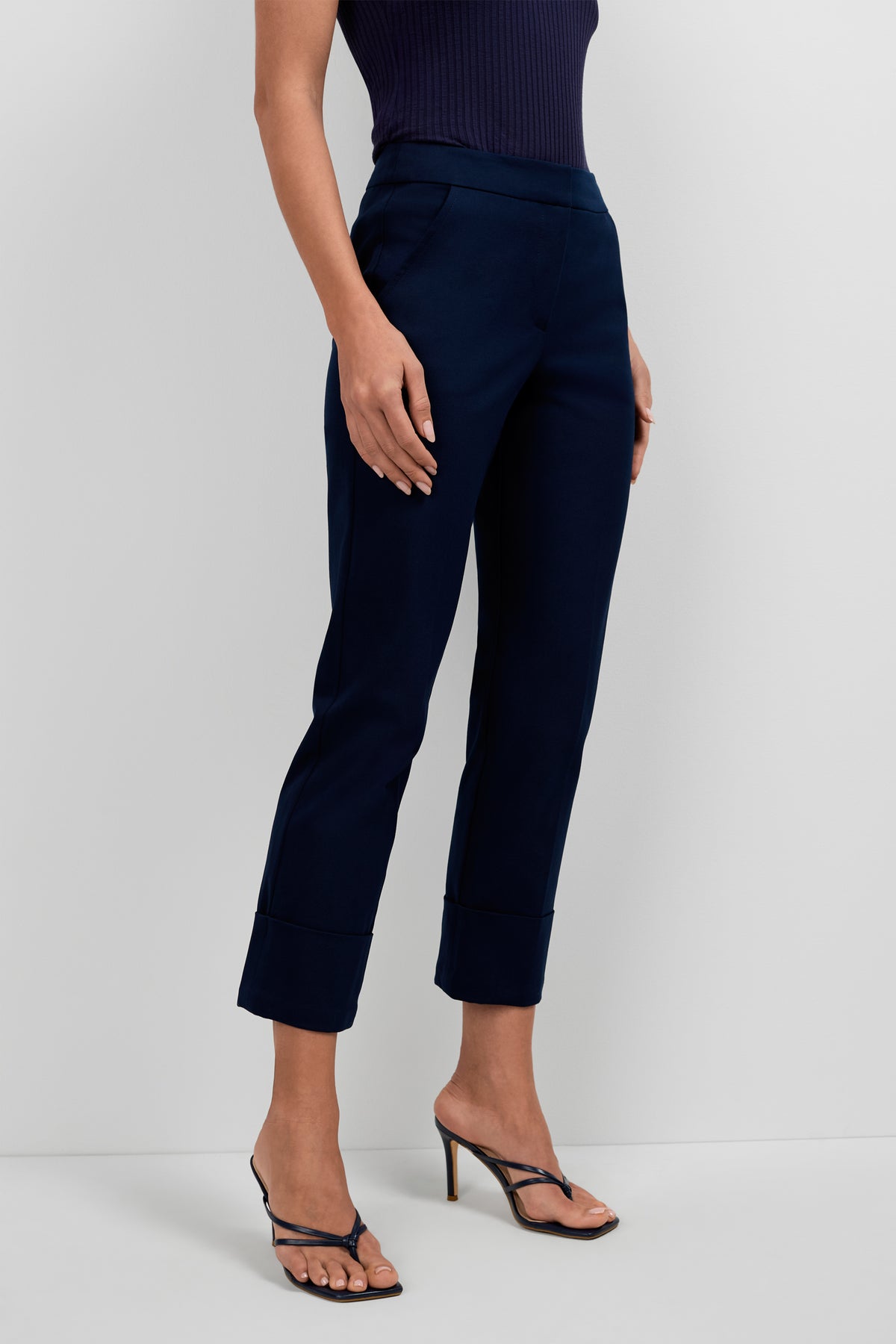 A person wearing Banshee Cropped Cuff Hem Pants in navy blue paired with black strappy high-heeled sandals, posing against a plain white background. Only the lower half of their body is visible.
