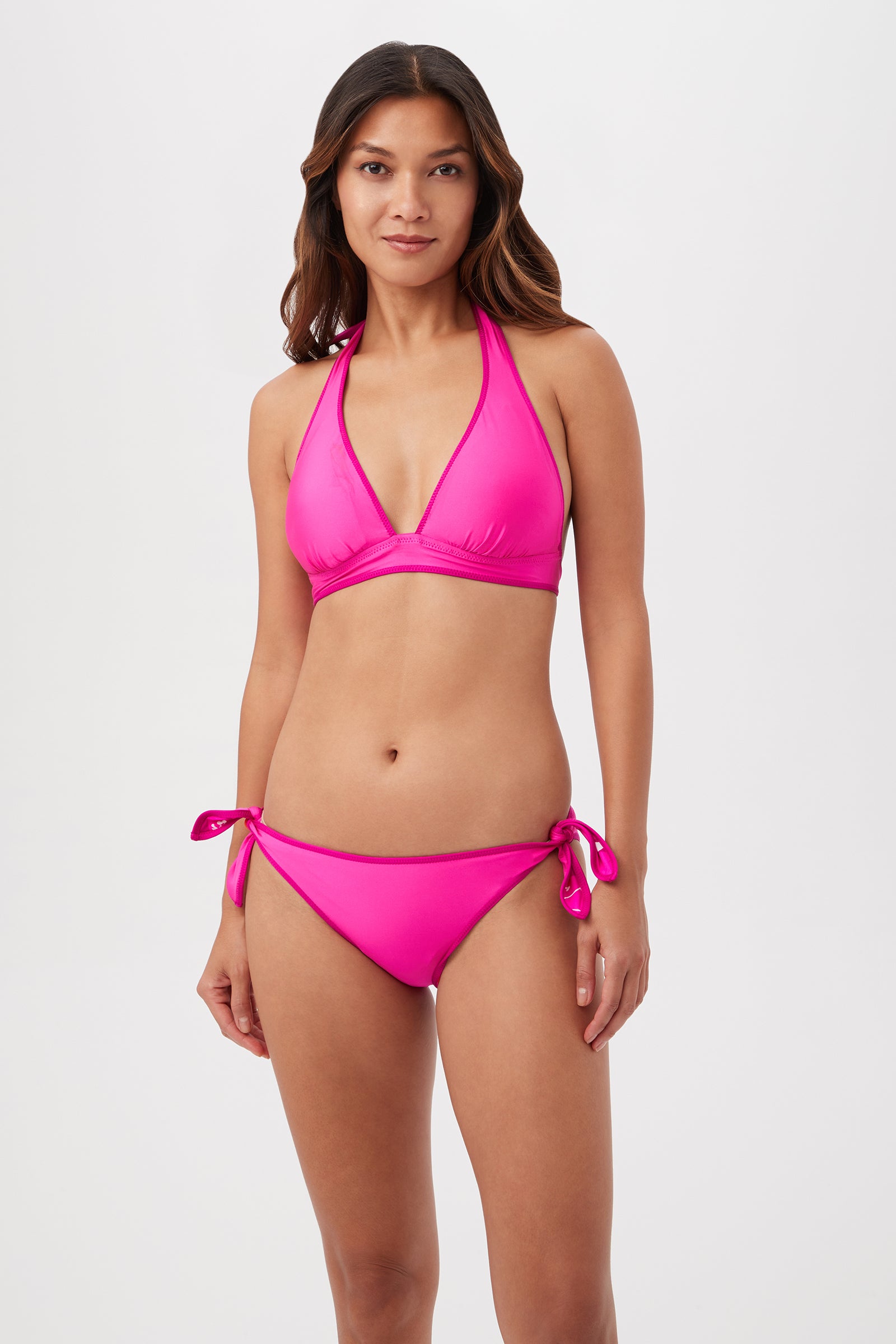 A woman poses against a white background, wearing the Trina Turk Bloom Halter Top Reverse bikini with tie-side bottoms. She has long brown hair, a neutral expression, and her arms rest naturally by her sides.