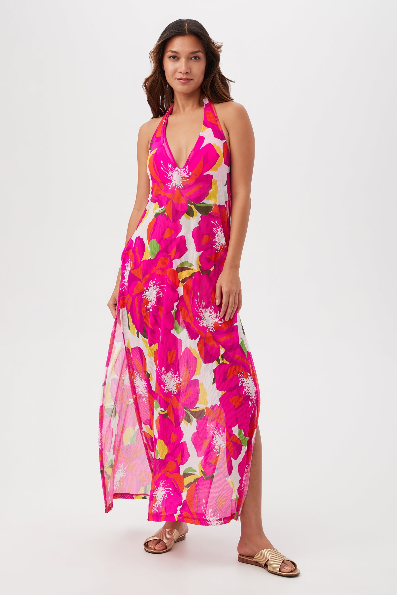 A woman stands against a plain white background wearing the Bloom Mesh Maxi Dress in a multi-color print with sleeveless, halter-neck design and side slits. She pairs it with tan flat sandals.