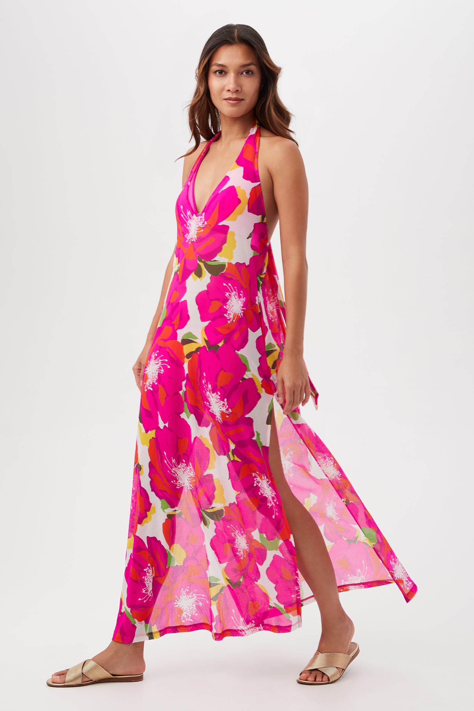 A woman stands against a white background wearing the Bloom Mesh Maxi Dress—a sleeveless, halter-neck maxi dress with a pink and red floral print and side slit—paired with beige slide sandals.