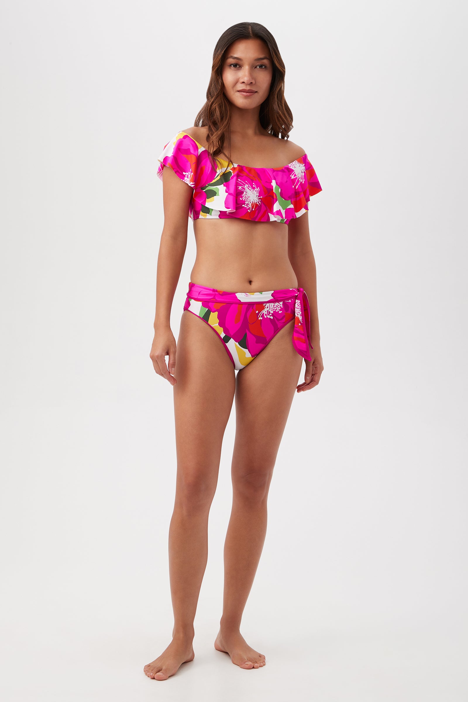 A woman stands barefoot against a white backdrop, wearing the Bloom Off The Shoulder Ruffle Top— a bright pink tropical print bikini with ruffled sleeves and high-waisted bottoms. She looks ahead with a relaxed expression.