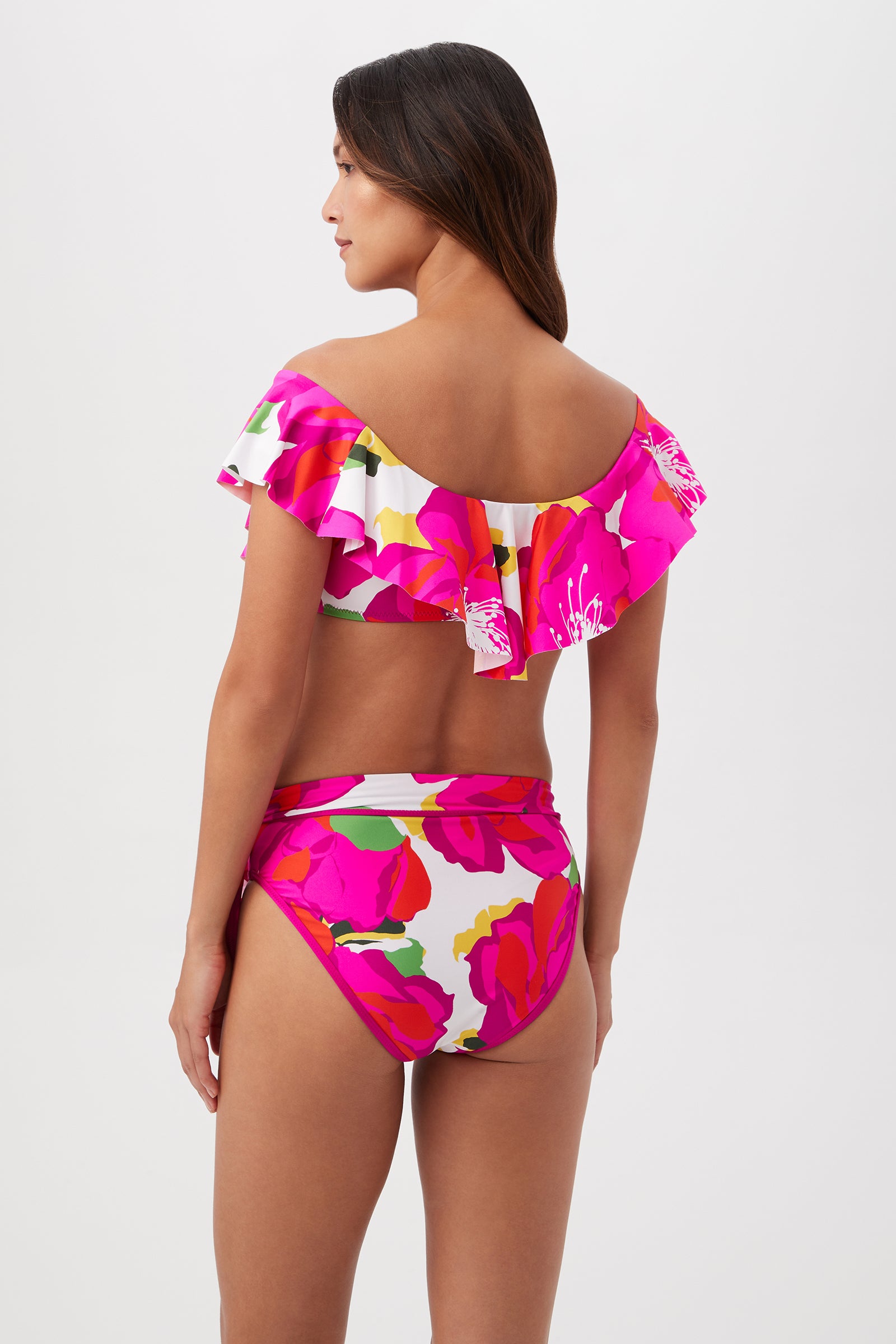 A woman with long brown hair stands facing away, wearing the Bloom Off The Shoulder Ruffle Top—a vibrant tropical print bikini with pink, red, and green patterns and ruffled sleeves—against a plain light background.