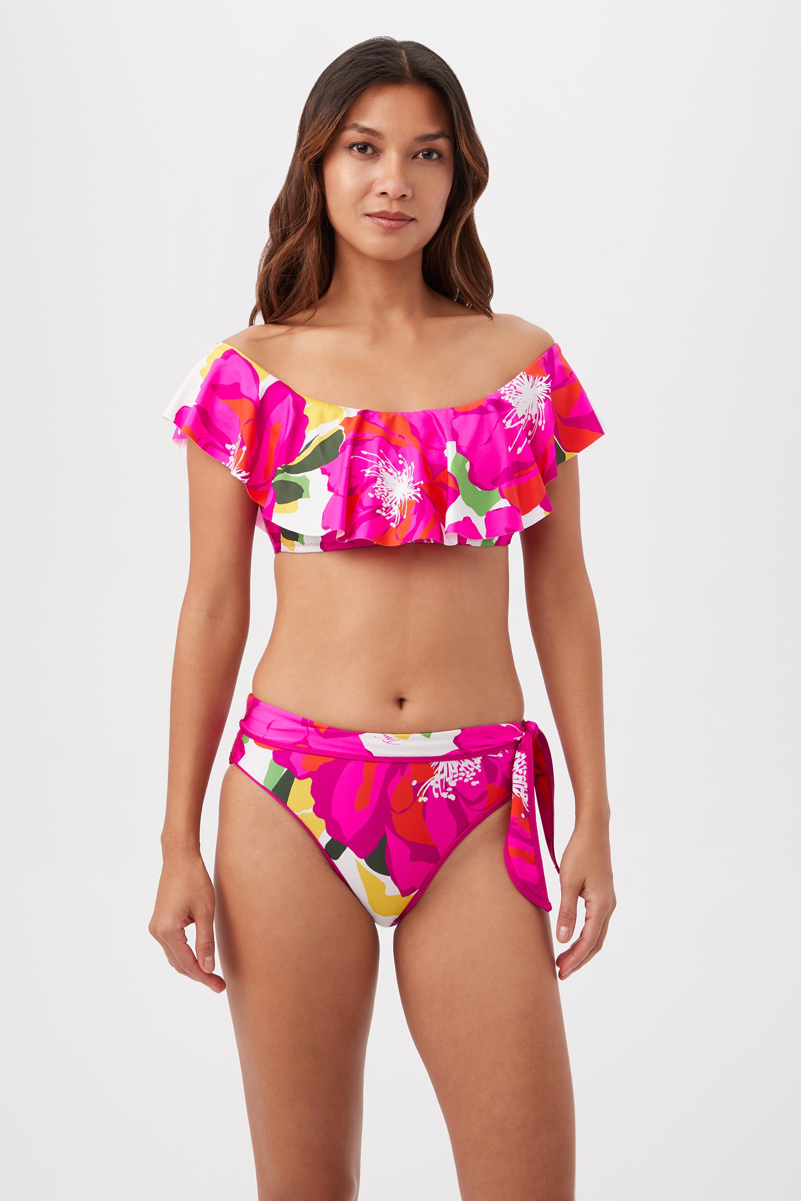 A woman with long brown hair wears the Bloom Off The Shoulder Ruffle Top in Multi, a bright tropical print swimwear set with a ruffle bandeau top and high-waisted side-tie bottoms featuring pink, white, and yellow florals.