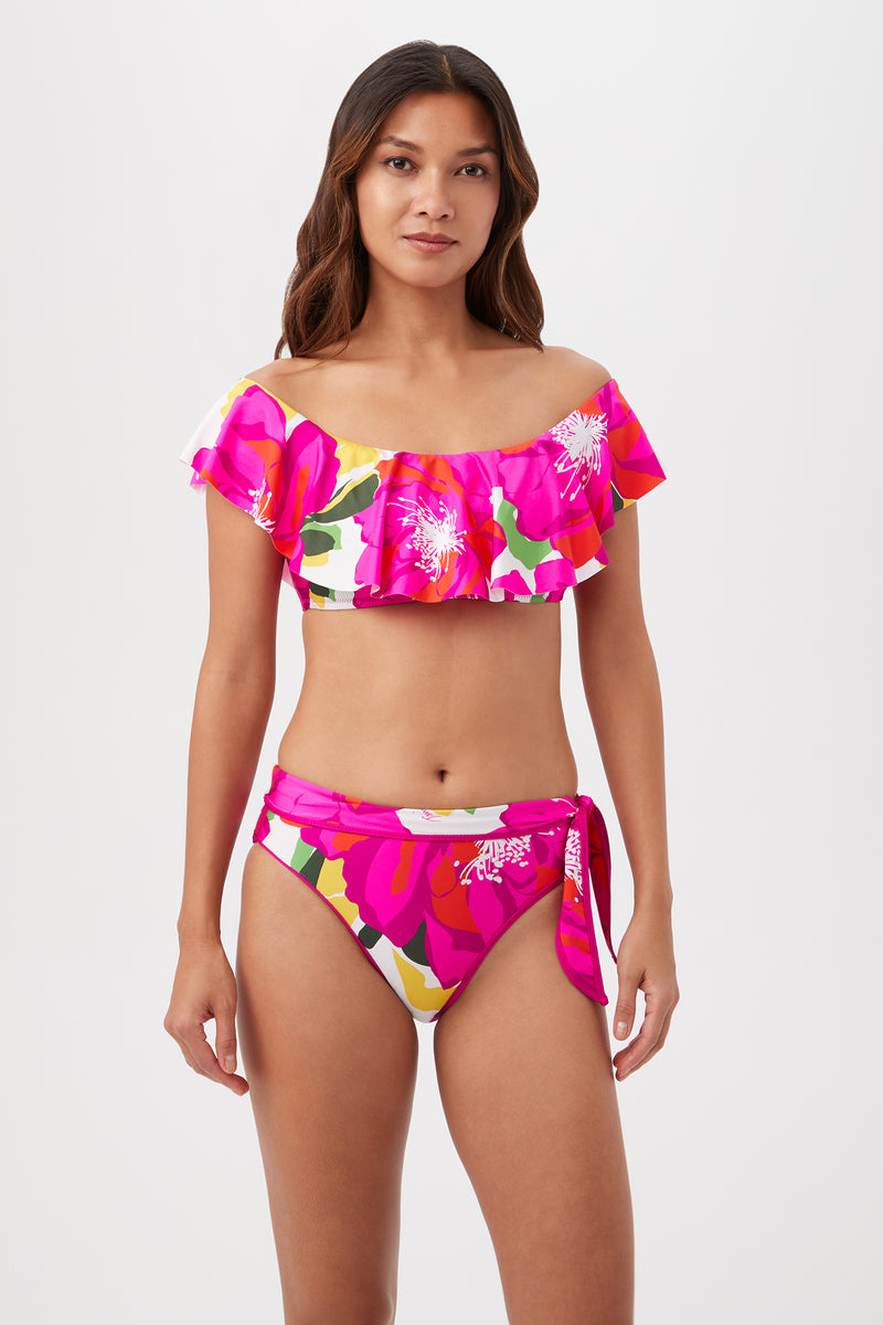 A woman with long brown hair wears the Bloom Off The Shoulder Ruffle Top in Multi, a bright tropical print swimwear set with a ruffle bandeau top and high-waisted side-tie bottoms featuring pink, white, and yellow florals.
