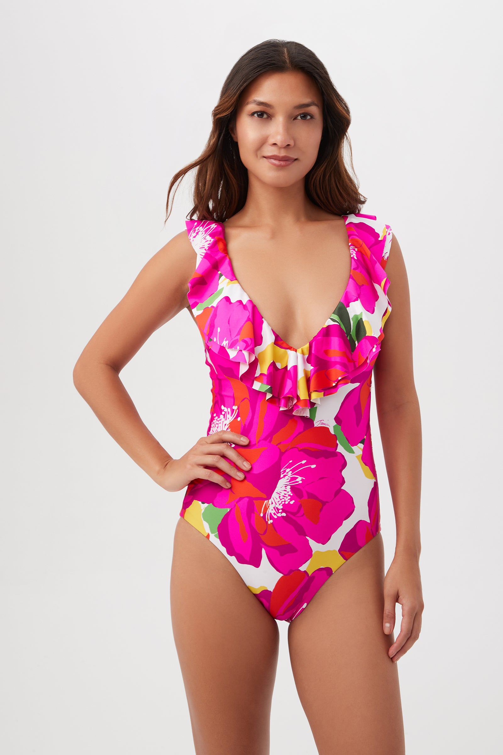 A woman stands against a plain white background wearing the Bloom Plunge Ruffle One Piece by Trina Turk in Multi, featuring a ruffle neckline and vibrant pink and red floral pattern.