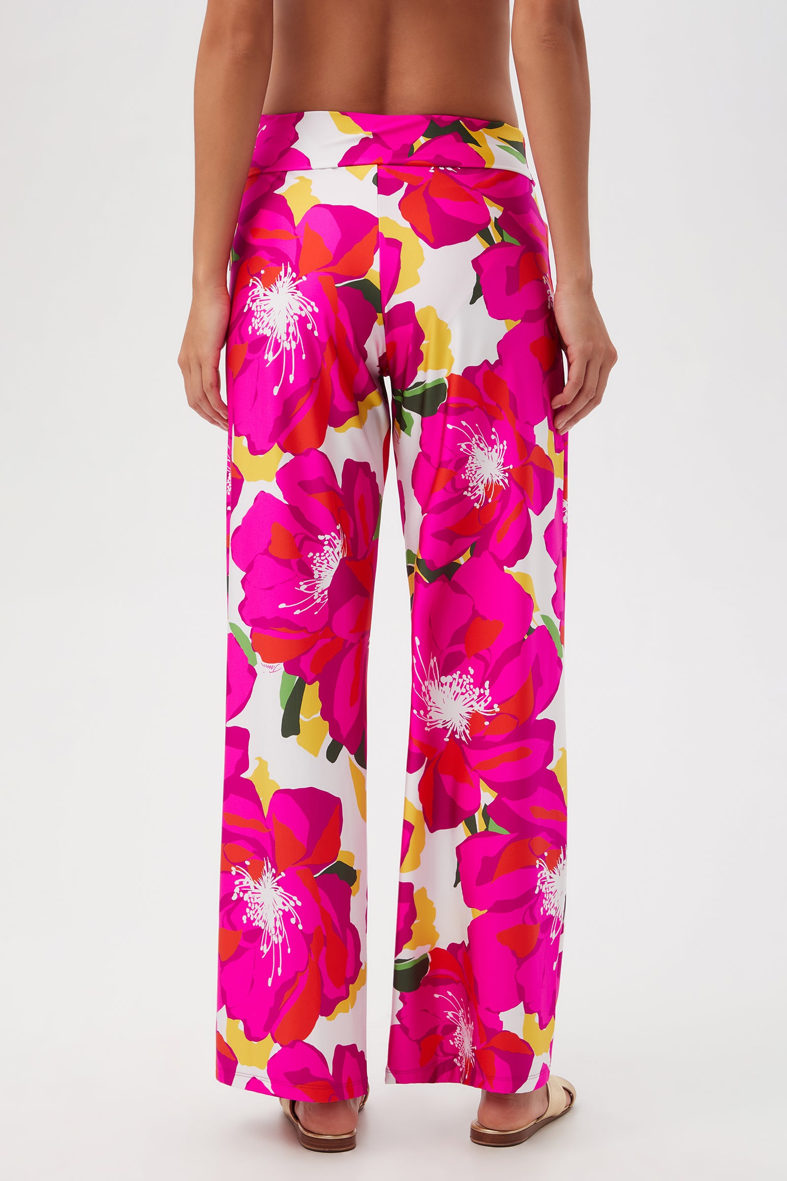 Model faces away, showcasing the Bloom Swim Pant—a stylish swim cover-up featuring a bold pink, yellow, and white floral pattern with wide legs. Only the lower back and legs are visible against a plain white background.