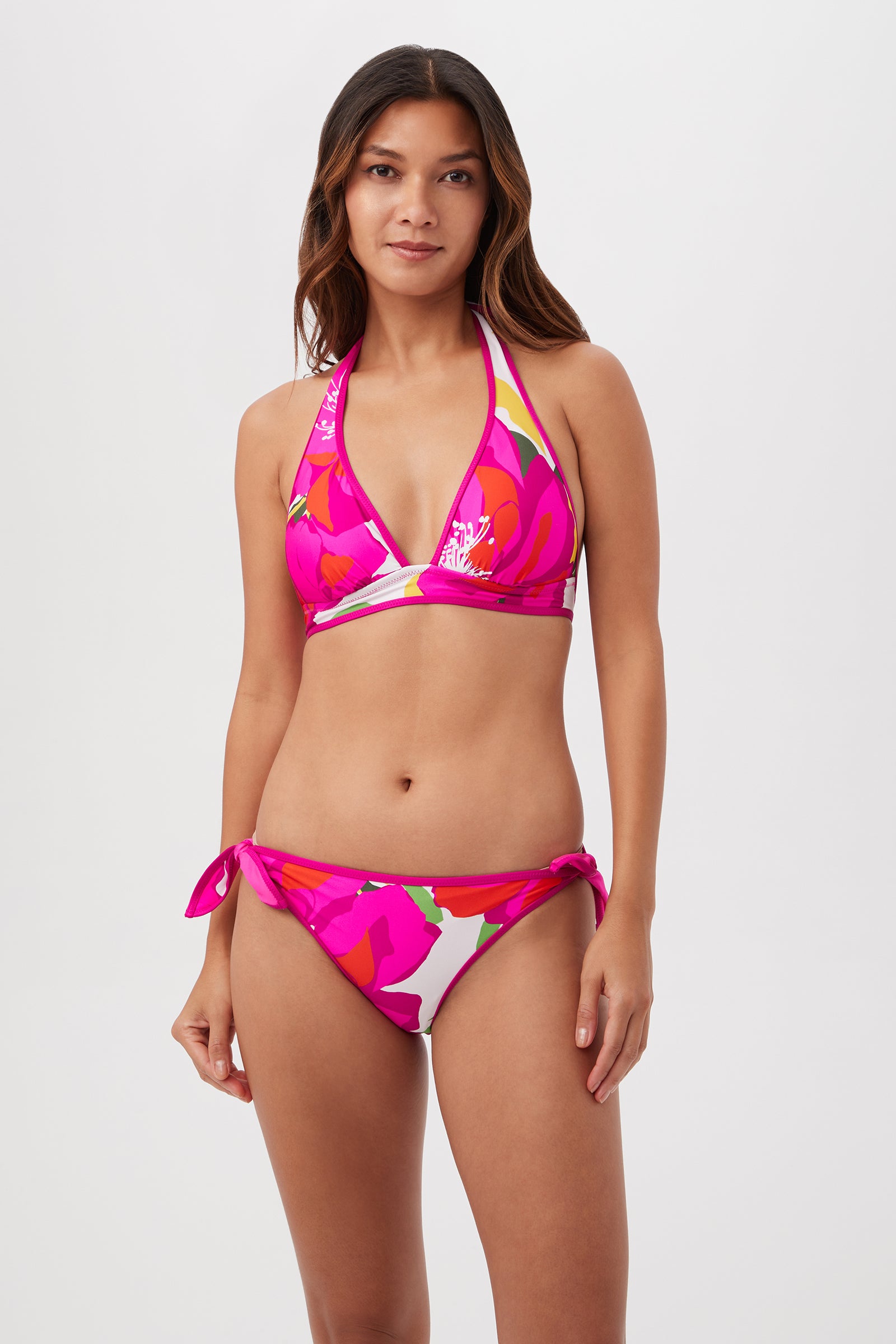 A woman stands against a plain white background wearing the Trina Turk Bloom Halter Top Reverse in Multi—a floral-patterned bikini with tie-side bottoms and a reversible halter top.
