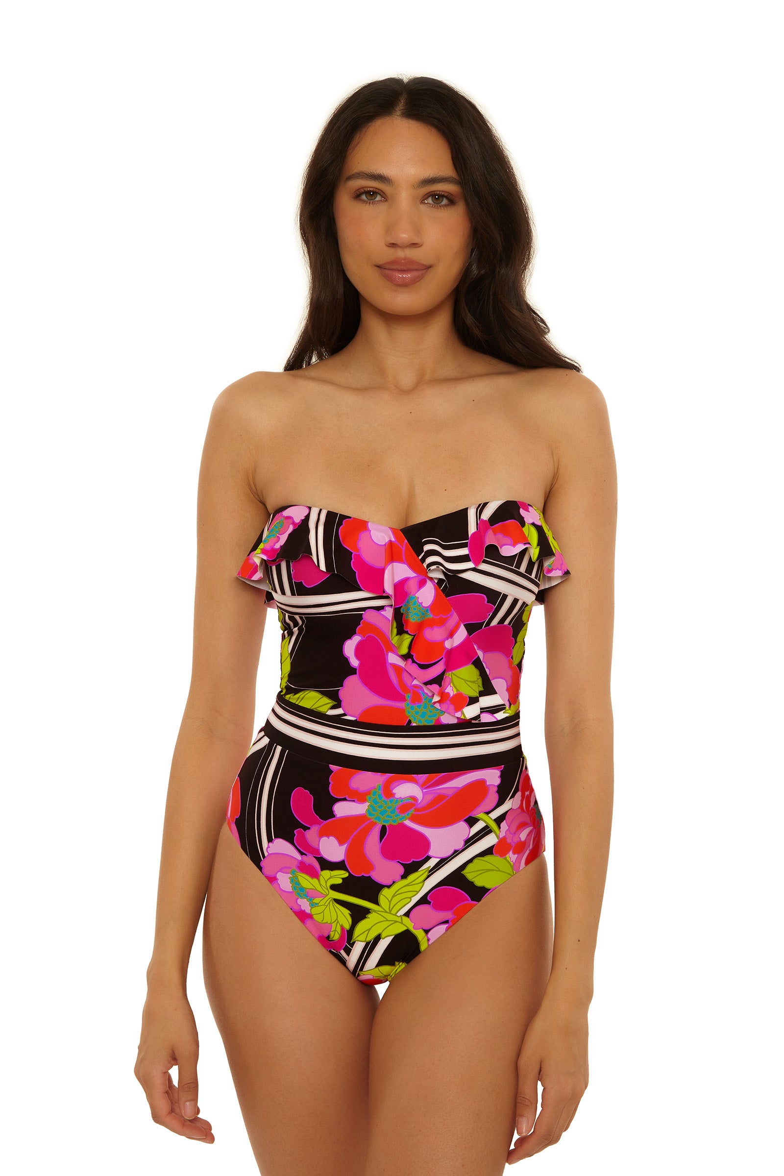 A woman stands facing the camera, wearing the Bandeau One-Piece Swimsuit in Bougainvillea Swim Knit, featuring a bold, colorful floral pattern on a black-and-white striped background.